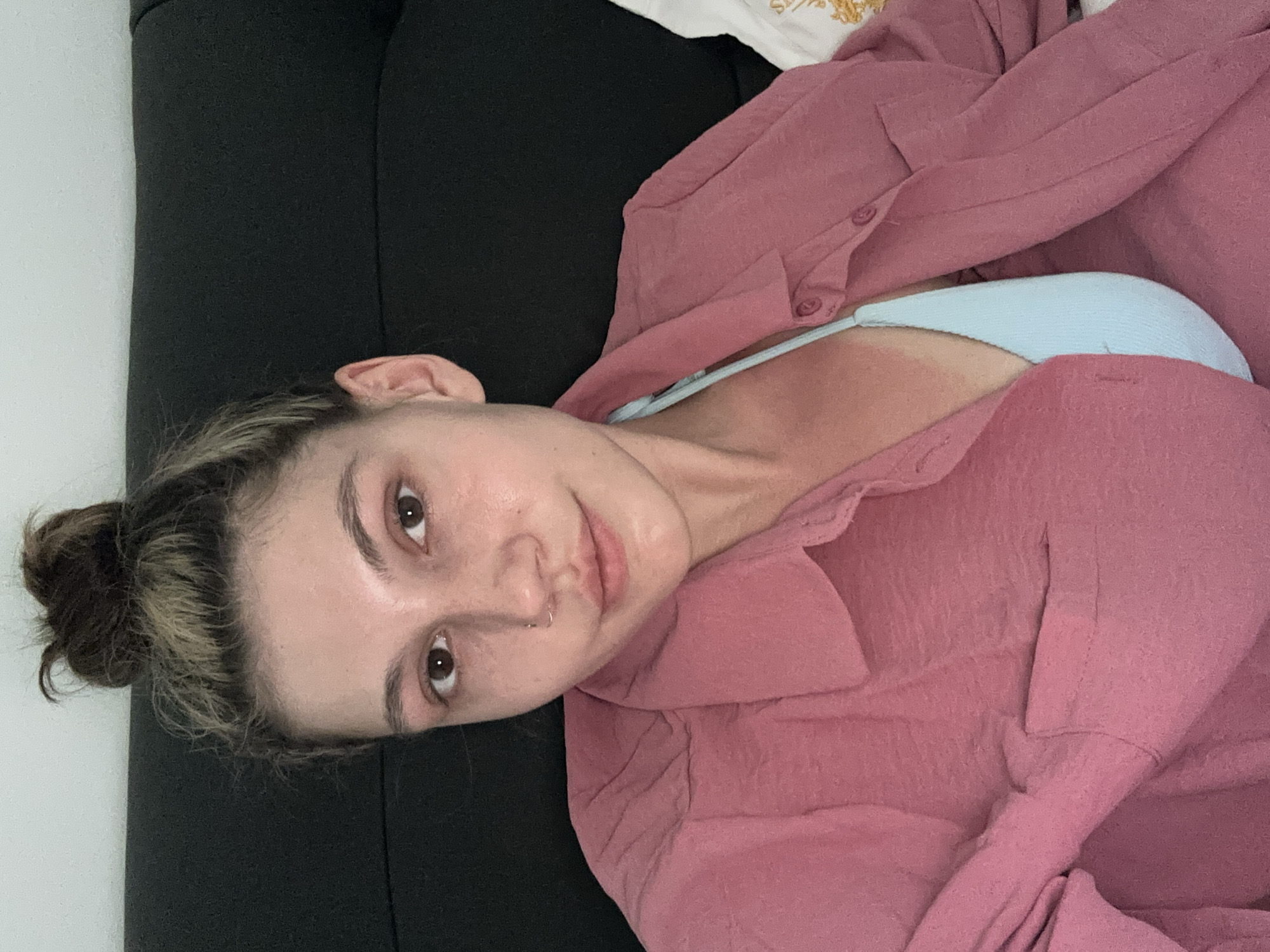 A woman with brown eyes and a nose piercing is lying on a dark surface, possibly a couch or bed, next to a white wall. She is wearing a light blue top under a light pink button-down shirt. Her hair is pulled back into a messy bun. The location of Baan Chaweng, Thailand is not discernible from the image.