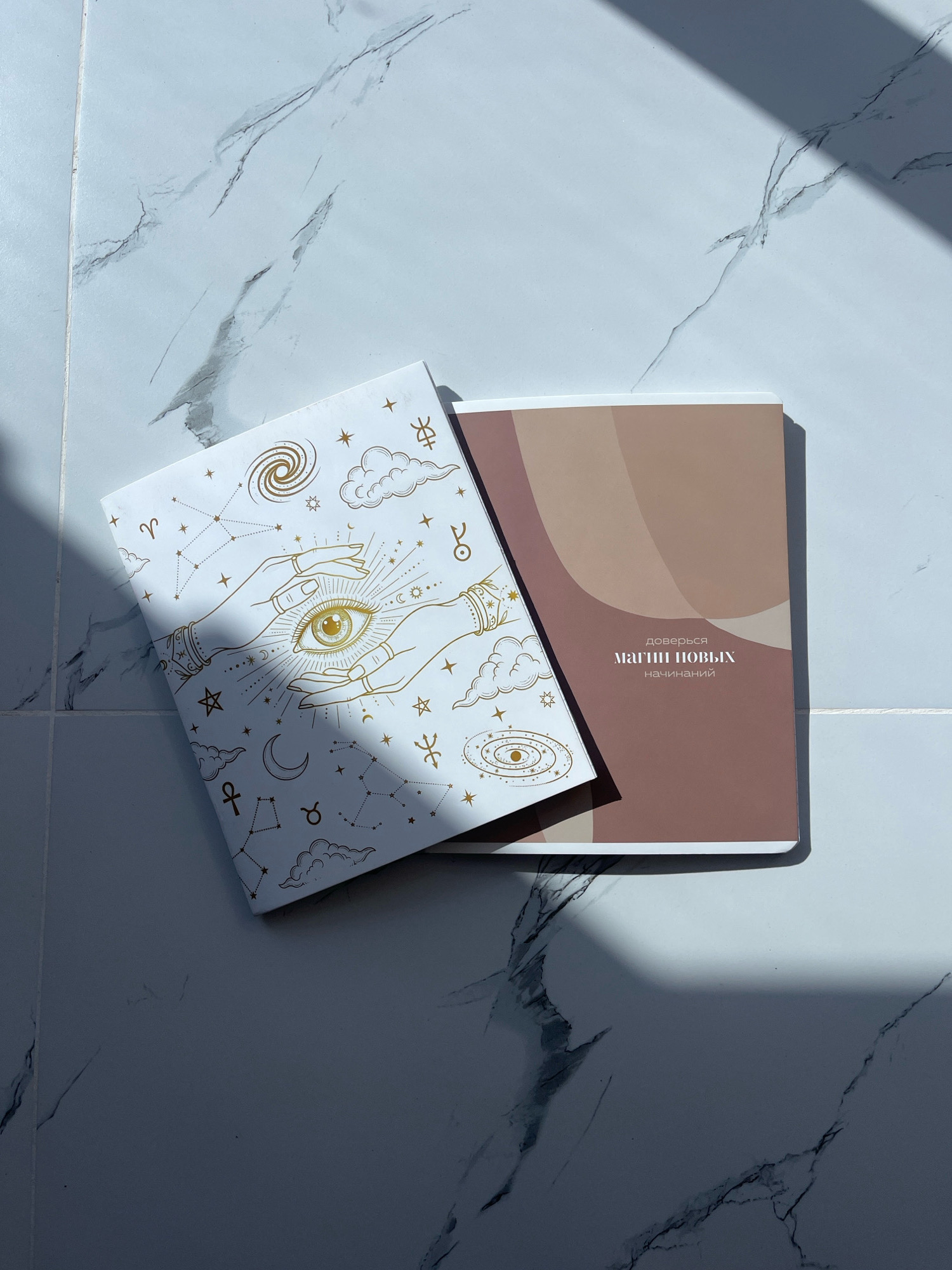Two notebooks are placed on a marble-patterned surface, illuminated by harsh overhead lighting casting dramatic shadows. The notebook on the left has a white cover adorned with gold celestial motifs, including hands holding an eye, constellations, stars, clouds, and zodiac symbols. The notebook on the right has a plain, muted brown cover with abstract, overlapping color blocks. Russian text is visible on the right notebook, reading "доверься магии новых начинаний" which translates to "trust the magic of new beginnings." The surface beneath the notebooks is a light-colored, marbled tile or countertop.