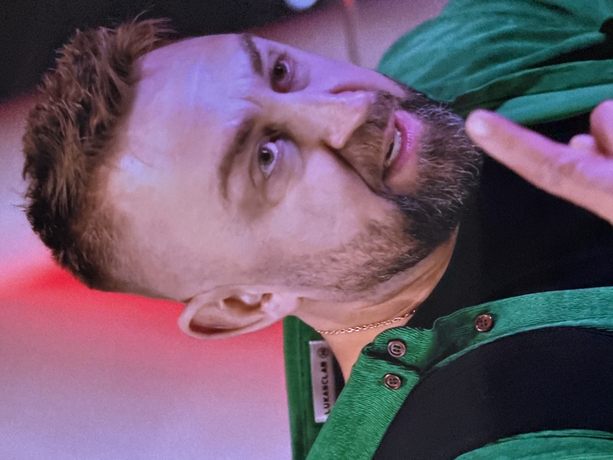 The image is a close-up shot of a man's face and upper torso, captured in what appears to be an indoor setting with colored lighting. The man has short, spiky brown hair, a beard, and is wearing a black shirt with a green corduroy-like jacket over it. The jacket has two visible buttons and a label that reads "LUKASCLAB". The background is out of focus and shows a blend of purple and red light, with a dark area on the right. The man's mouth is slightly open, as if he is speaking or reacting to something, and he is pointing with his right index finger towards the viewer. The lighting creates a dramatic effect on his face. The image does not provide any explicit visual cues to determine its specific location within Moscow, Russia.