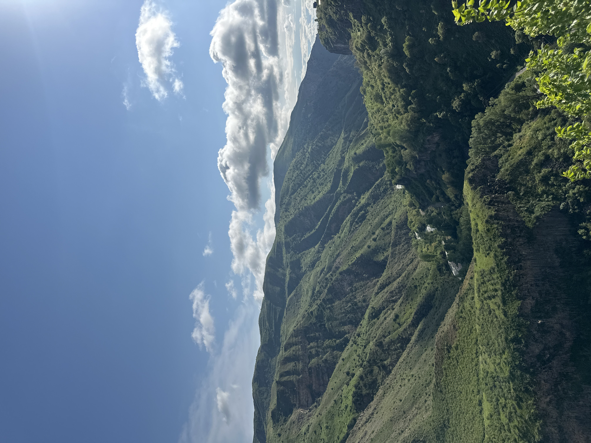 This is a landscape photograph taken near the city of Garni, Armenia. It depicts a steep, green mountain slope under a partly cloudy sky. The mountain is covered in vegetation, with some rocky outcrops visible. The sky is blue with scattered white clouds, and the sun is shining, casting shadows on the mountain. There are no people visible in the image.