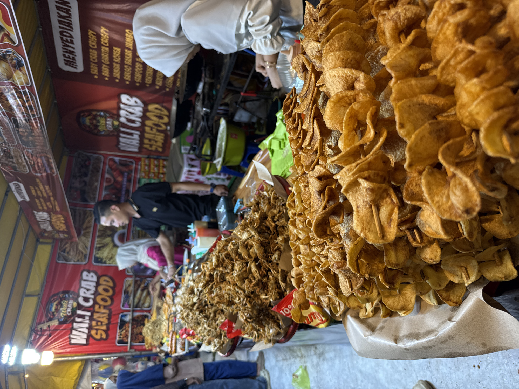The image depicts a food stall in Geulumpangsulu Timu, Indonesia. The stall, named "Wali Crab Seafood," offers a variety of seafood dishes including baby crab crispy, baby gurita crispy, jabar korea, and bakso bakar jumbo. A man wearing a black shirt is behind the counter, presumably preparing food. A person wearing a white hijab is also visible near the counter. In the foreground, there are stacks of spiral-cut potatoes on skewers, with some promotional packaging, likely advertising discounts.