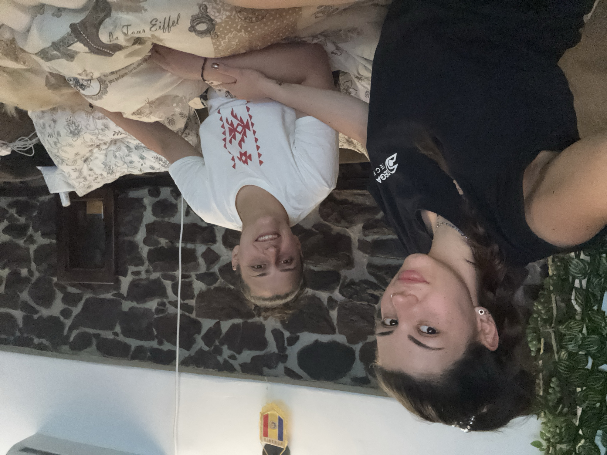 Two young women are indoors, lying on a bed, in what appears to be a room with a stone wall and a decorative flag. The woman on the left is wearing a white t-shirt with red geometric designs and holding hands with the other woman. The woman on the right is wearing a black t-shirt. The bed is covered with patterned bedding, featuring the Eiffel Tower design. Plants are visible on the right edge of the frame. The decorative flag has the colors blue, yellow, and red. The city where the photo was taken is Belopoptsi, Bulgaria.