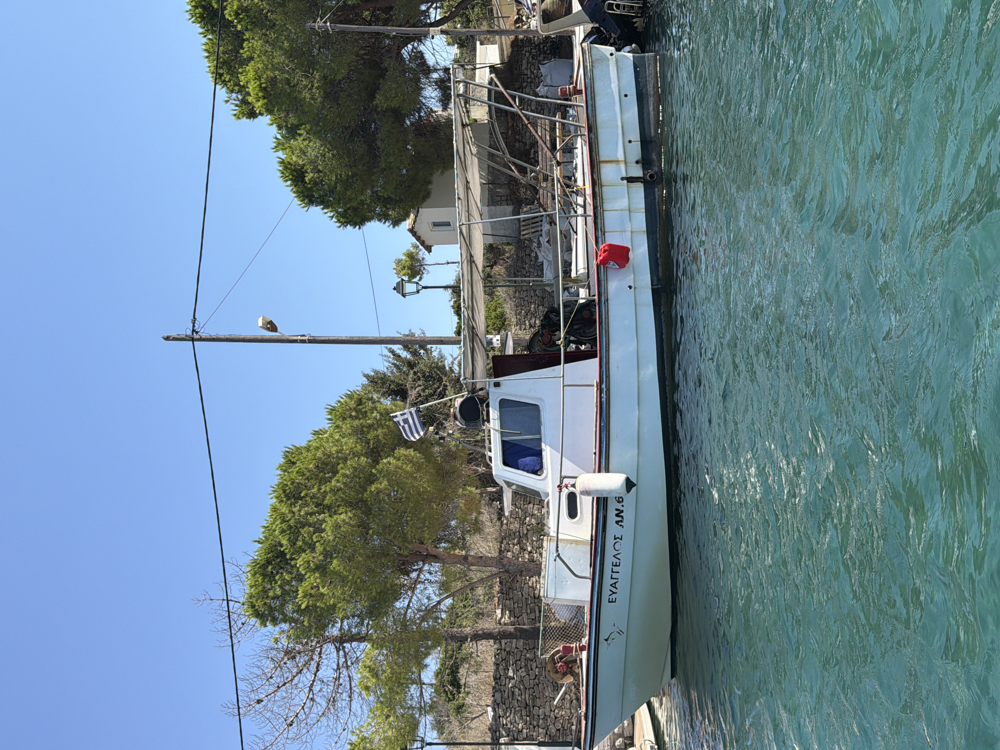 A white fishing boat with the name "ΕΥΑΓΓΕΛΟΣ ΑΝ.6" written in black on its hull is docked in the clear turquoise water. The boat has a white cabin with blue-tinted windows and a canvas canopy supported by metal poles. A Greek flag is flown from a mast at the stern. The background shows a stone wall, lush green trees, and a small white building with a red-tiled roof. A lamppost with a classic design stands on the shore. The scene is set under a clear blue sky, suggesting a sunny day in the Municipal Unit of Kalamos, Greece. The water is calm with gentle ripples. No people are visible in the frame.