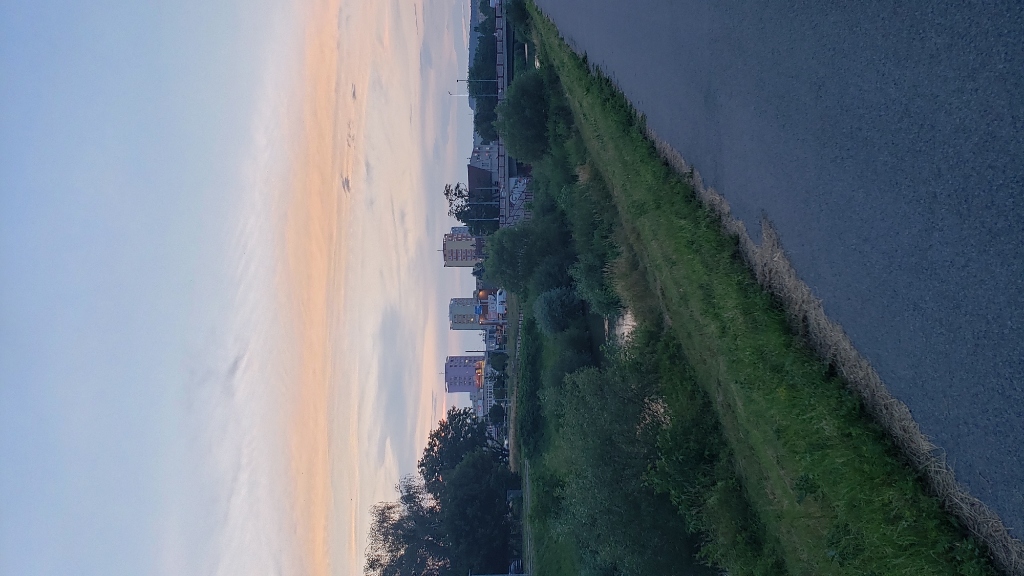 This is a vertical, medium-distance, eye-level, outdoor shot of a picturesque sunset over the city of Prešov, Slovakia. The sky is a blend of soft pink, orange, and pale blue hues. In the distance, a line of modern, colorful apartment buildings stands tall against the horizon, some of them illuminated. A bridge spans across a calm body of water, possibly a river, with lush green trees and dense bushes lining its banks. A paved pathway or road runs along the right side of the frame, bordered by a strip of tall grass. The overall scene evokes a sense of tranquility and urban beauty at dusk.