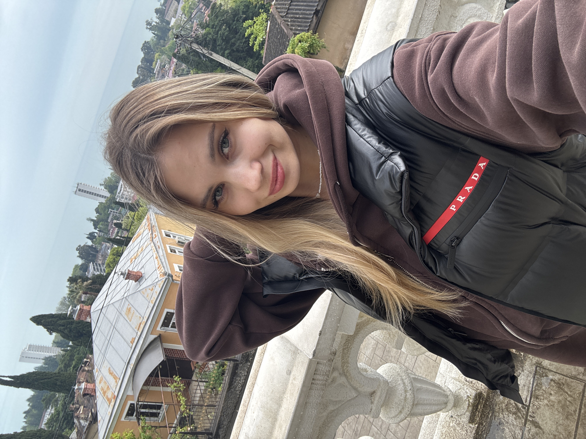 A young woman is taking a selfie on a balcony with decorative stone balustrades. She has long blonde hair, fair skin, and is wearing a brown hoodie under a black puffer jacket with a "PRADA" logo. She is smiling slightly.
In the background, a cityscape stretches towards a body of water. The buildings consist of residential homes with grey and orange roofs. Trees are scattered throughout the cityscape. In the far background, a tall white building is visible next to the body of water.
This scene is likely located in Sokhumi, Abkhazia.