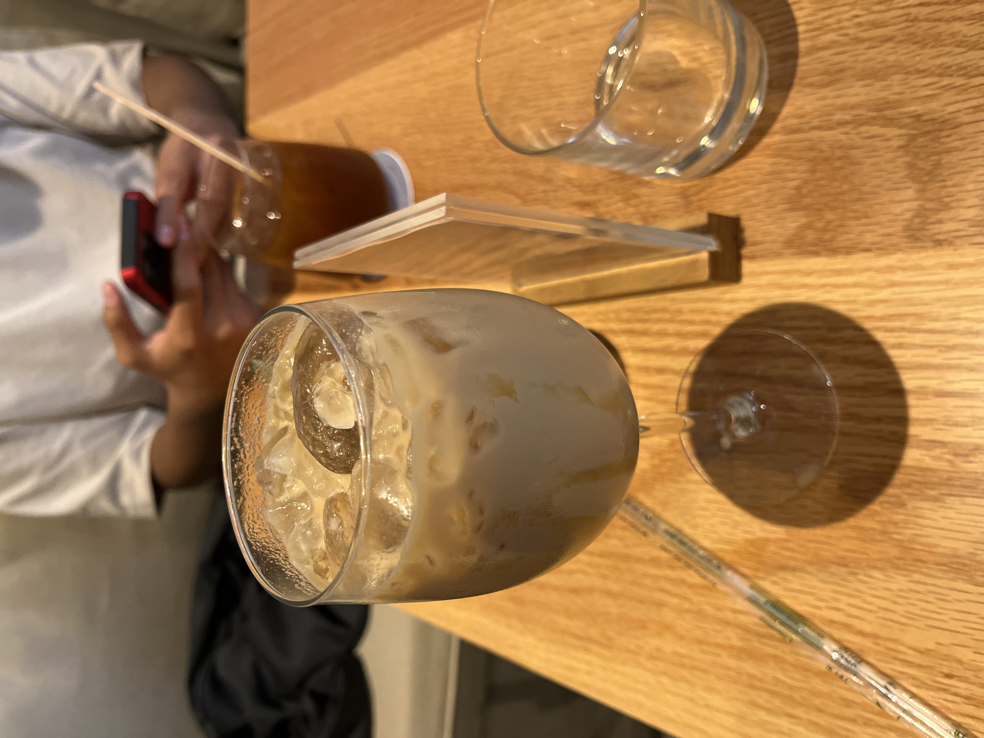The image captures an overhead view of a light-colored wooden table at what appears to be a cafe or restaurant in San Bernardino Tlaxcalancingo, Mexico. The scene features multiple beverages and a partially visible person.
On the left side, a person wearing a light-colored top is seen holding a red smartphone with both hands. Adjacent to their hands is a glass filled with a clear, dark amber liquid, presumably iced tea, containing a thin, light-colored straw.
Towards the right and in the foreground, a stemmed glass holds a creamy, light brown iced beverage, likely an iced latte or coffee, brimming with ice cubes. Its tilted angle offers a clear view of the drink's texture. Further back on the right, an empty clear drinking glass is visible.
Between the iced tea glass and the empty glass, a small, clear acrylic stand is present on the table, seemingly holding a blank white card or sign. A long, transparent straw with subtle green markings extends across the bottom right section of the table.
The lighting is soft, creating distinct shadows from the glasses on the wooden surface, indicating an indoor setting. The overall impression is one of a casual and relaxed gathering. No discernible text is visible within the frame.