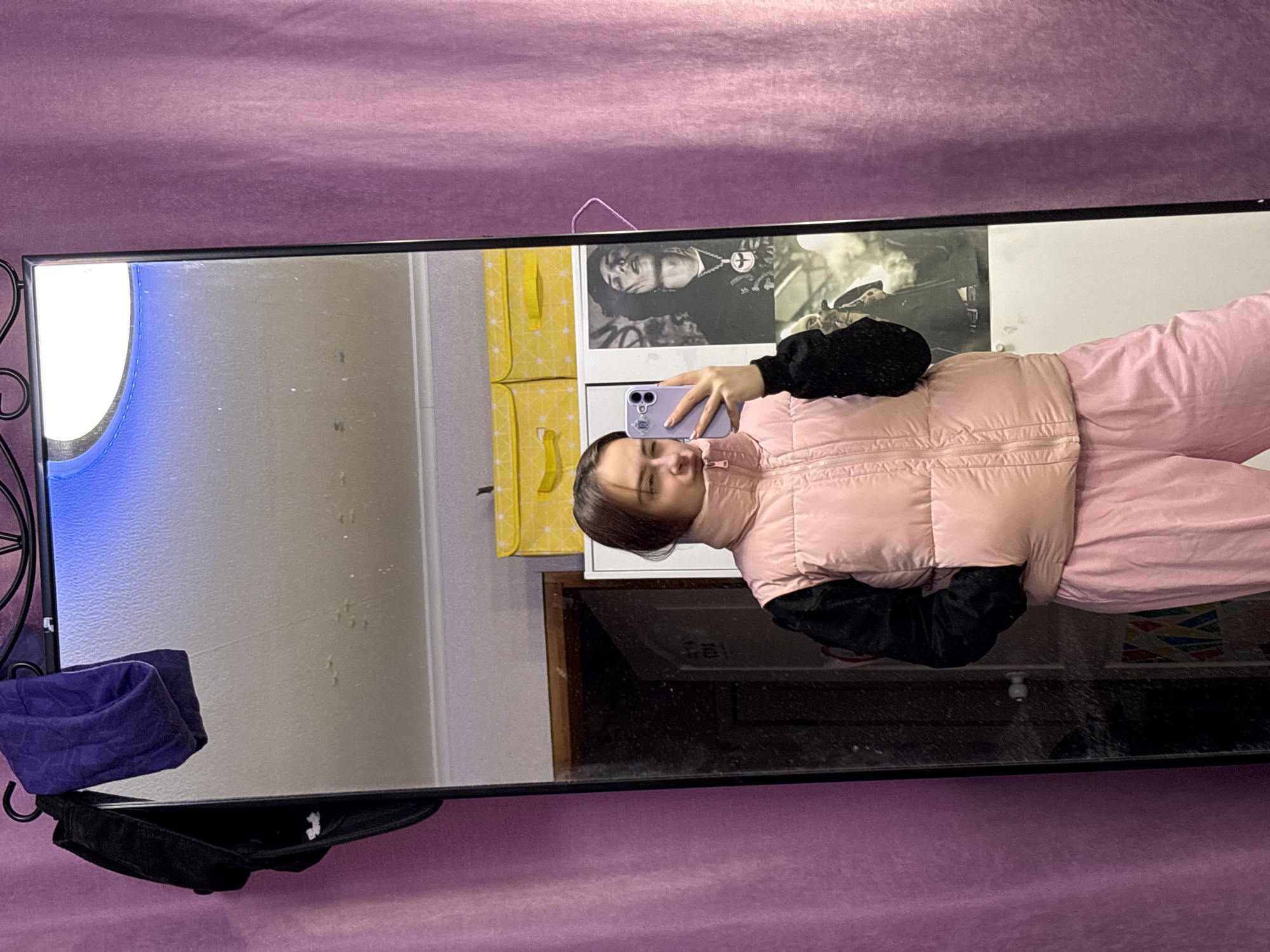 A young woman is taking a selfie in a mirror. She is wearing a pink quilted vest over a black long-sleeved shirt and matching pink pants. The mirror is hanging on a purple wall. The reflection in the mirror shows a room with yellow storage boxes and a framed photograph of a musician. There are also a black cap and a folded purple garment hanging on the mirror's frame. The location is likely in Прикамский, Russia, as indicated in the instructions.