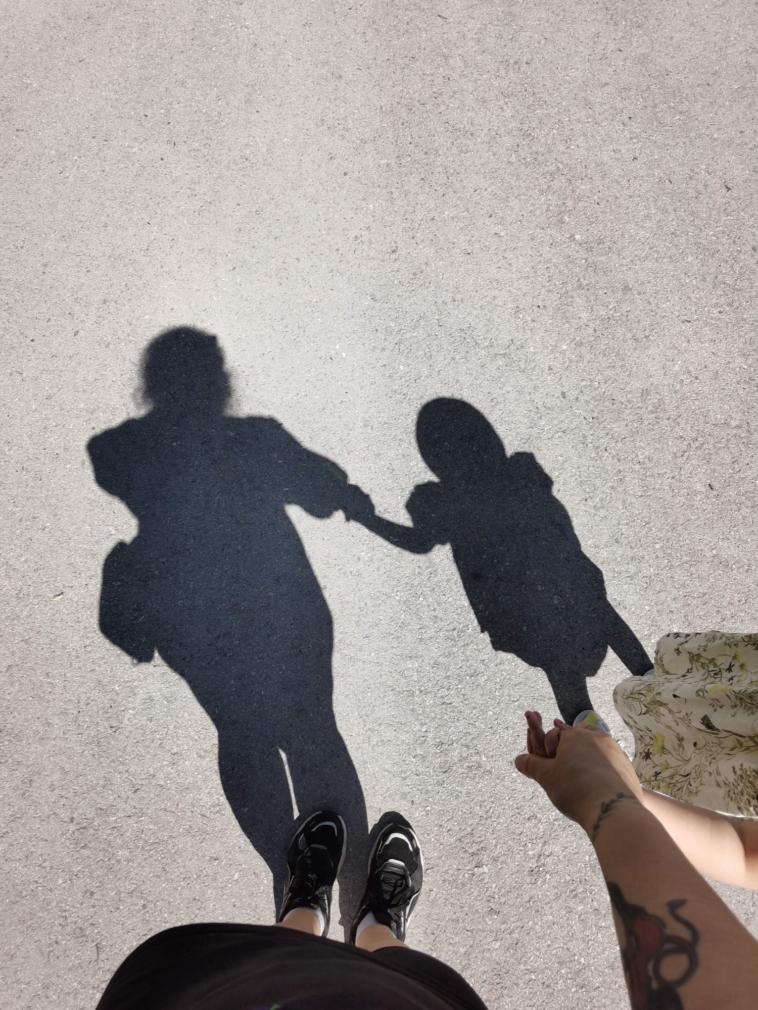 The image depicts an aerial view of a person wearing black athletic shoes and a black skirt, and a second person wearing a floral patterned skirt. They are standing on a light gray asphalt surface. The shadows of the two figures are cast on the asphalt, indicating a bright sunlight source. The shadows suggest one is an adult holding hands with a child. The tattooed arm and hand of the adult person are visible in the lower right corner, clasping the hand of the child figure.
While I can describe the scene, I cannot determine the location in the city of Perm, Russia, from the visual data provided.