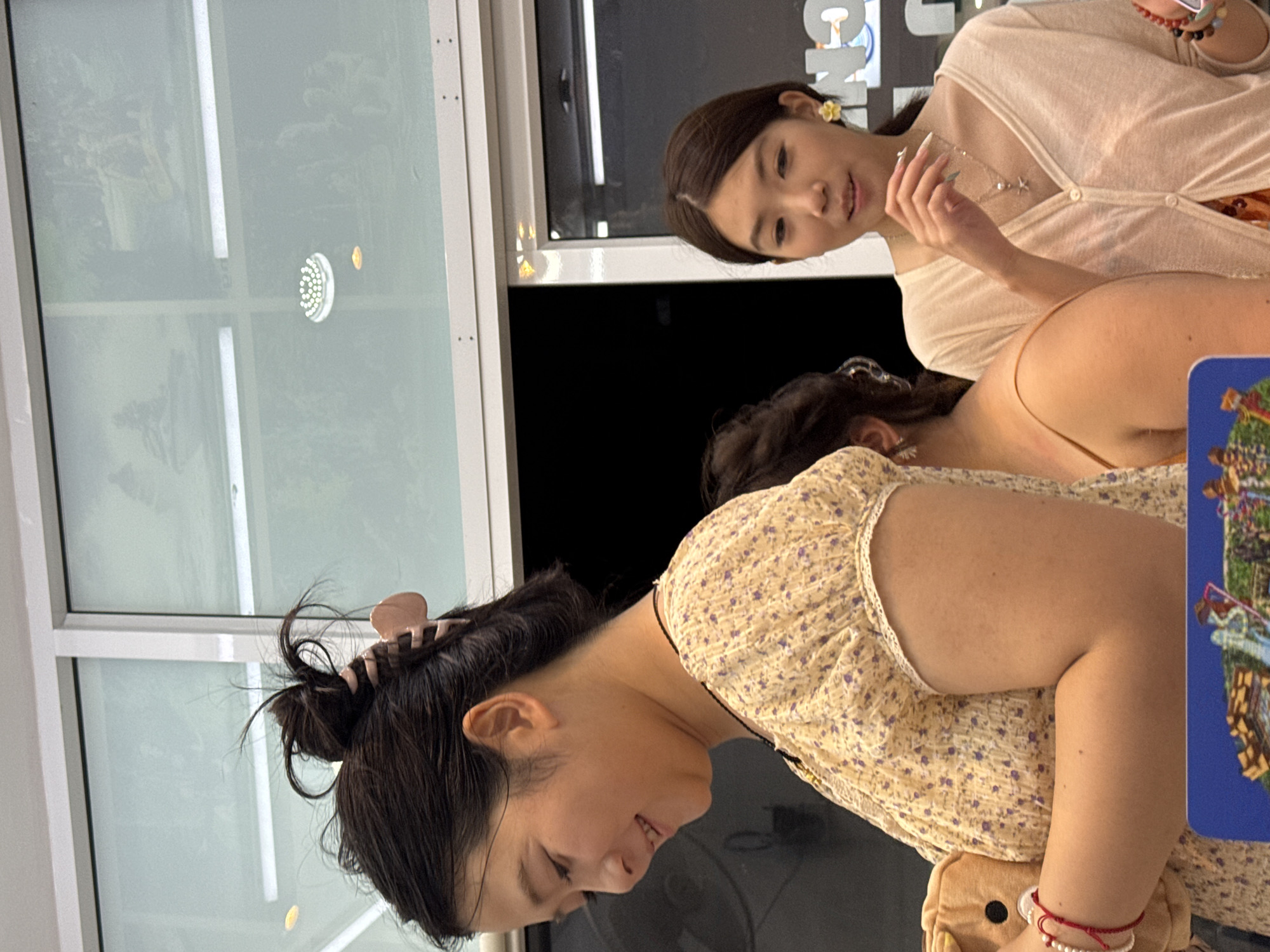 The image shows two young Asian women indoors, likely in a commercial setting. The woman on the left, with dark hair pulled up in a bun secured by a pink clip, wears a yellow floral print blouse and is holding a small plush toy. She has a red string bracelet on her left wrist. The woman on the right, with dark hair down, wears a pale pink cardigan over a light-colored outfit and has a yellow flower earring. She is looking to her left and appears to be interacting with someone off-camera. Behind them, large windows reveal reflections and potentially images or displays, with overhead lighting visible. The environment appears to be a shop or store.