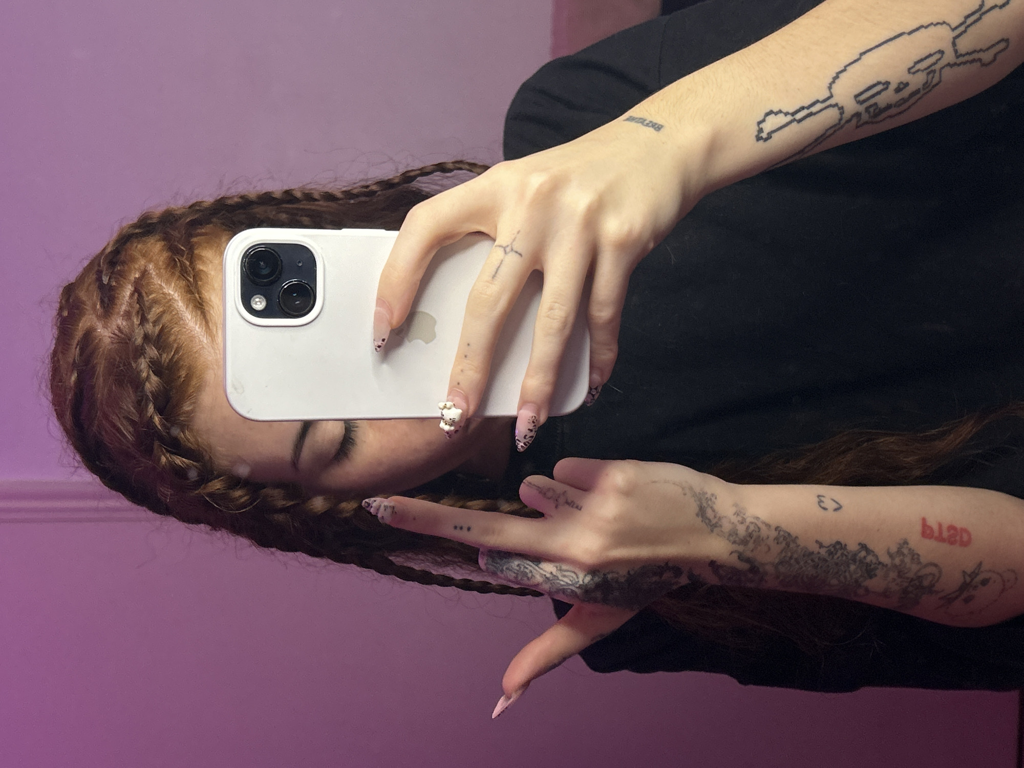 A person with long, braided reddish-brown hair is holding a white smartphone in their right hand, taking a selfie. The person is wearing a black shirt and has multiple tattoos on their arms and hands. The background is a plain light purple wall. The person's eyes are closed. The phone has three camera lenses and an Apple logo on the back. The fingers of the person's left hand are making a peace sign. The person has long, decorated fingernails. The location is not discernable from the image.