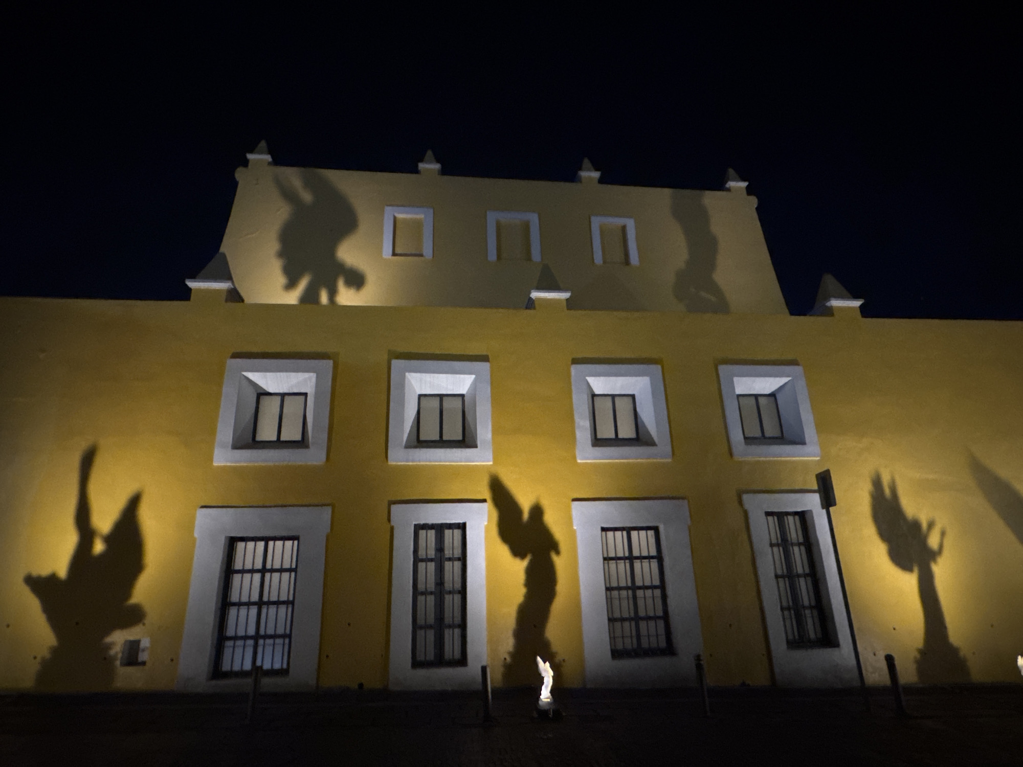 The image depicts a yellow building at night in the city of Puebla, Mexico. The building has a flat facade with rows of windows and rectangular decorative elements on the roof. Shadows of figures with wings are projected onto the facade, appearing to be the shadows of statues or figures positioned in front of the building. The sky is dark, and the light source illuminating the building appears to be at ground level, casting the shadows upward. The ground in front of the building is dark and partially obscured.