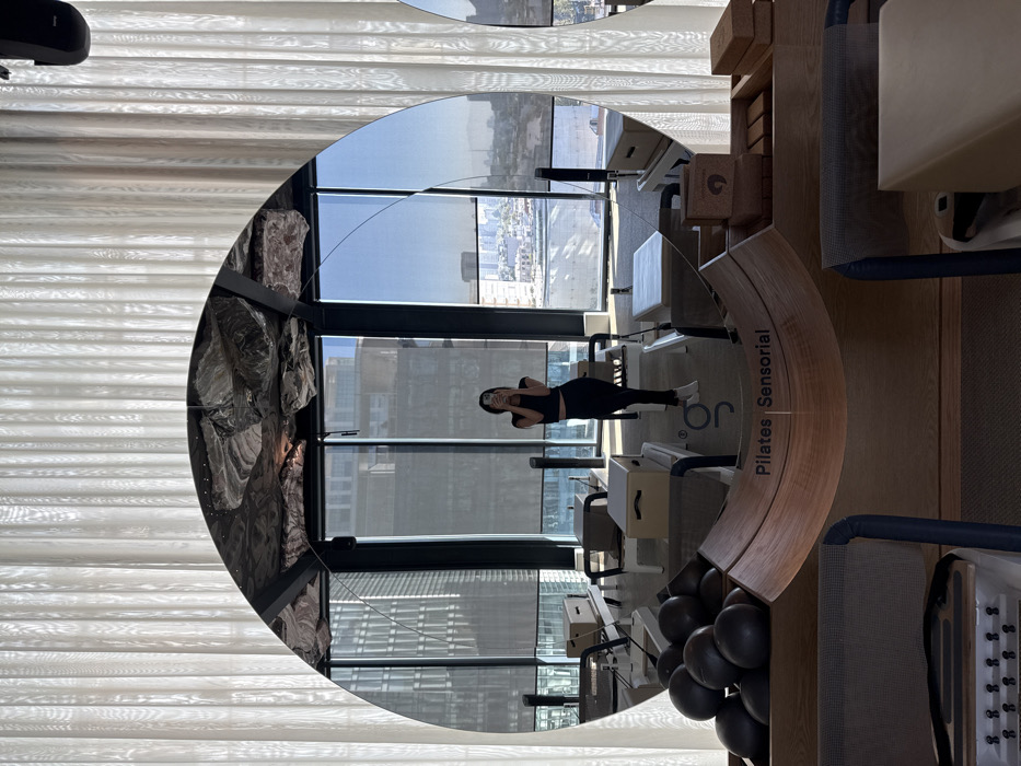 A round mirror reflects a pilates studio. The mirror shows a woman in workout clothes in the center. Pilates equipment, including reformers and wooden elements labeled "Pilates Sensorial," are visible. A cityscape is seen through the window. The left side of the mirror reflects a textured wall, while the right shows exercise equipment and black pilates balls. Curtains are seen in the upper-left corner.