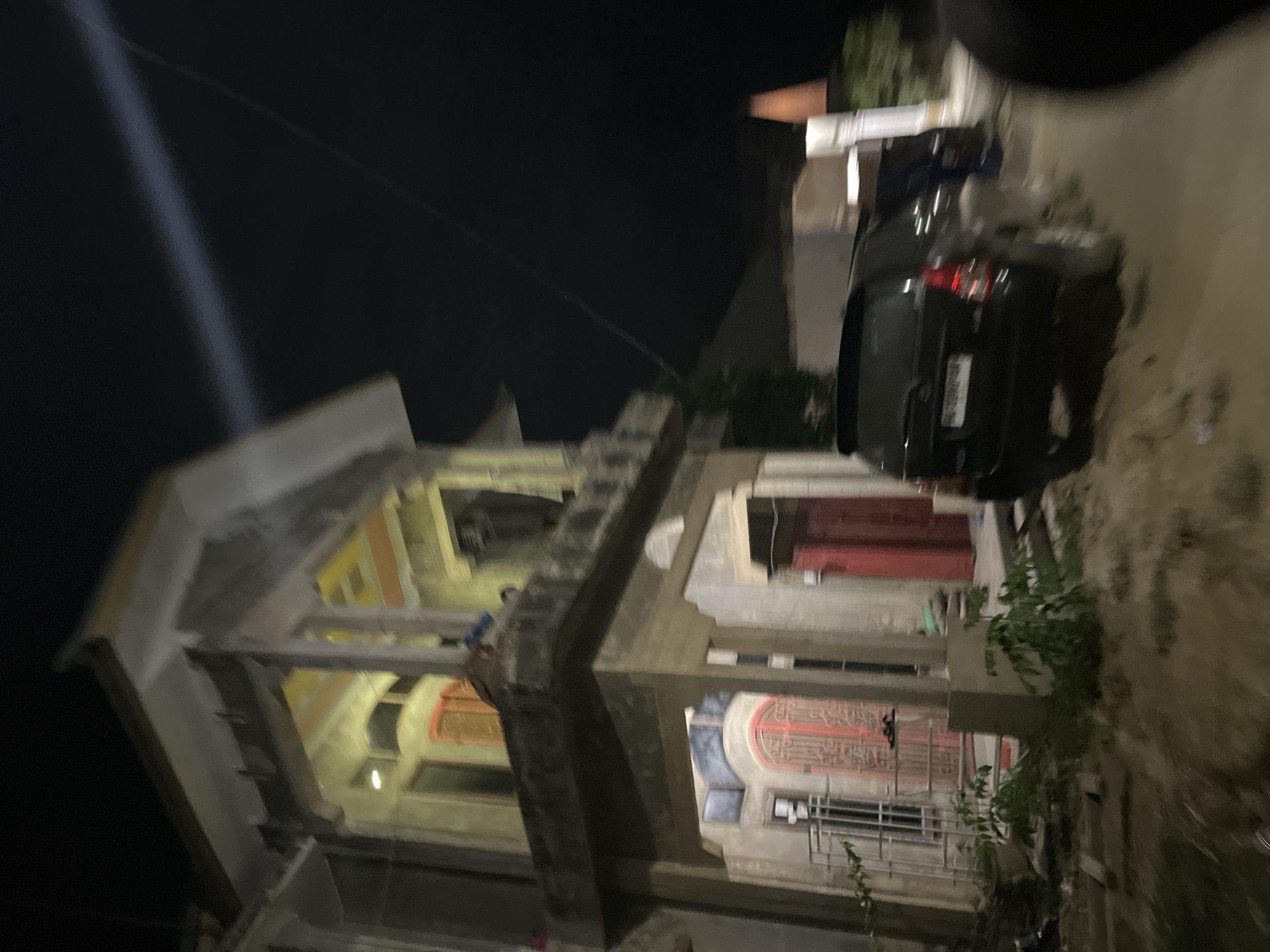 The image depicts a street scene at night in Menanggini, Indonesia, likely during fair weather. The focus is on a multi-story building with intricate architectural details, featuring arched windows with decorative grilles and balconies. The building appears to be illuminated from within, casting a warm glow on its facade. In front of the building, a dark-colored SUV is parked, its taillights visible. A person is seen on one of the balconies, possibly observing the surroundings or engaging in an activity. The street surface is wet, suggesting recent rain or a street cleaning. The overall atmosphere is quiet and residential, with the darkness of the night punctuated by artificial lights. The image is captured from a slightly elevated angle, offering a clear view of the building's structure and the parked vehicle. No visible text is present in the image.