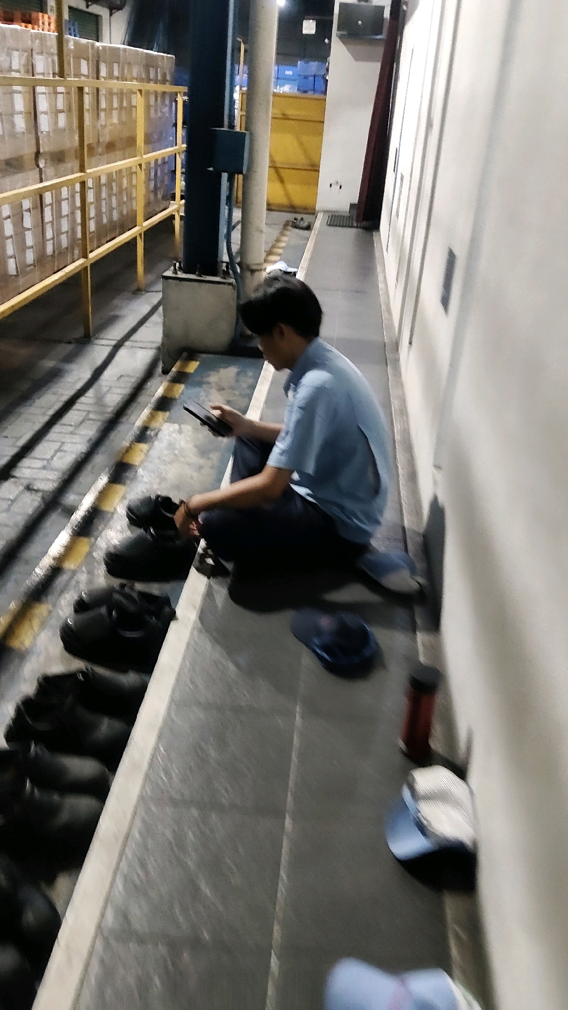 A young man is squatting on a raised concrete ledge in what appears to be a warehouse or factory setting. He is wearing a light blue short-sleeved collared shirt and dark pants, and he is looking down at a black smartphone he is holding in his hands. Beside him, on the concrete ledge, is a pair of black shoes. Further down the ledge, there are several more pairs of black shoes lined up. To his right, on the floor, is a dark blue cap and a red cylindrical object, possibly a thermos or bottle. In the background, to the left, stacks of boxes are visible behind yellow guardrails. The environment suggests a working or storage area. The man appears to be on a break or resting. The location is identified as Mekarmukti, Indonesia.