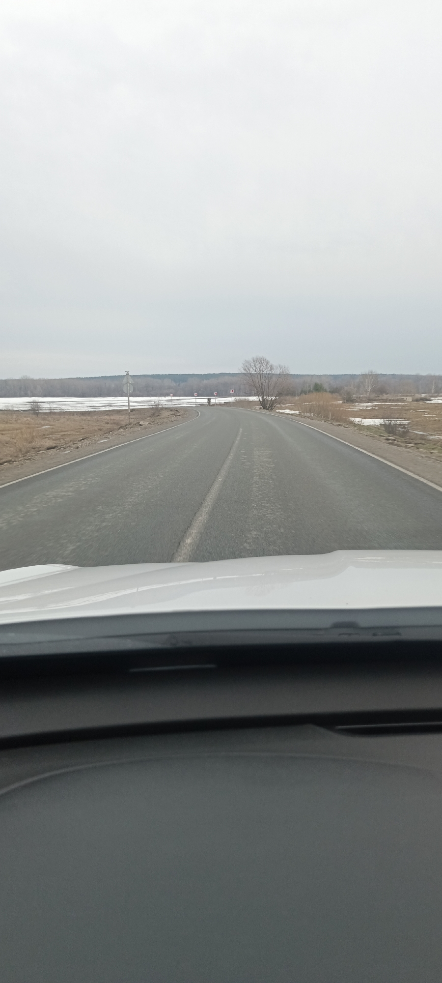 The image depicts a view from inside a vehicle, looking out onto a straight stretch of asphalt road. A single white dividing line runs down the center of the road. The road is flanked on either side by bare, dry land, interspersed with patches of snow or ice. A line of trees and shrubs is visible in the distance. The sky is overcast and grey. The vehicle's dashboard and part of the hood are visible in the foreground. The location appears to be a rural or semi-rural area.