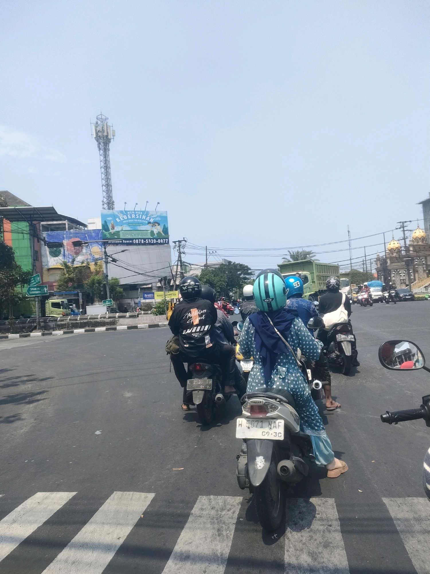 The image captures a street scene in Surabaya, Indonesia, likely an intersection, indicated by the presence of a pedestrian crossing and multiple vehicles. A queue of motorcycles is stopped at the crossing, suggesting traffic. On the right side of the frame, a gold-domed building, possibly a mosque, is visible in the background. In the mid-ground, a large billboard with text in Indonesian, promoting cleanliness, is mounted on a building. A cellular tower is prominently featured against the bright, clear sky. The people on the motorcycles are wearing helmets, and one rider in the foreground is wearing a blue and white polka-dot outfit. The environment appears to be urban, with buildings and utility poles lining the street.