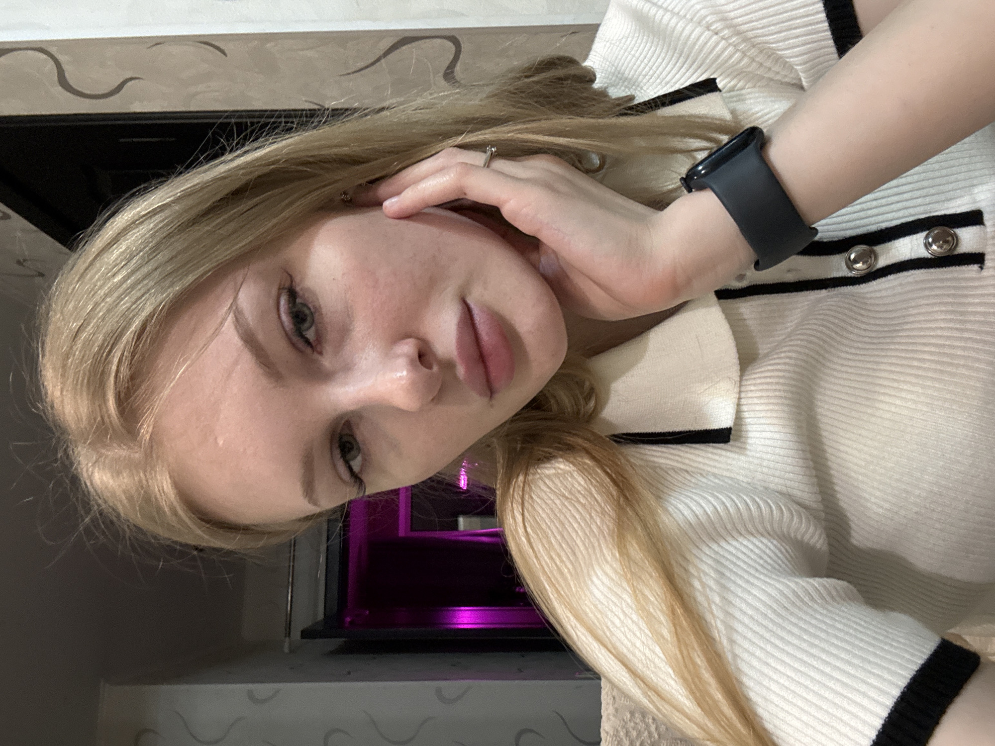 A fair-skinned young woman with blond hair is seen in a close-up shot, likely a selfie. She has green eyes and pink lips. Her right hand is near her face. A black smartwatch is on her left wrist. She wears a white, ribbed top with black trim and silver buttons. A silver ring is on one of the fingers of her right hand. In the background is a dark object illuminated with purple light and a patterned wall. The location is not specified in the image and therefore cannot be determined.
