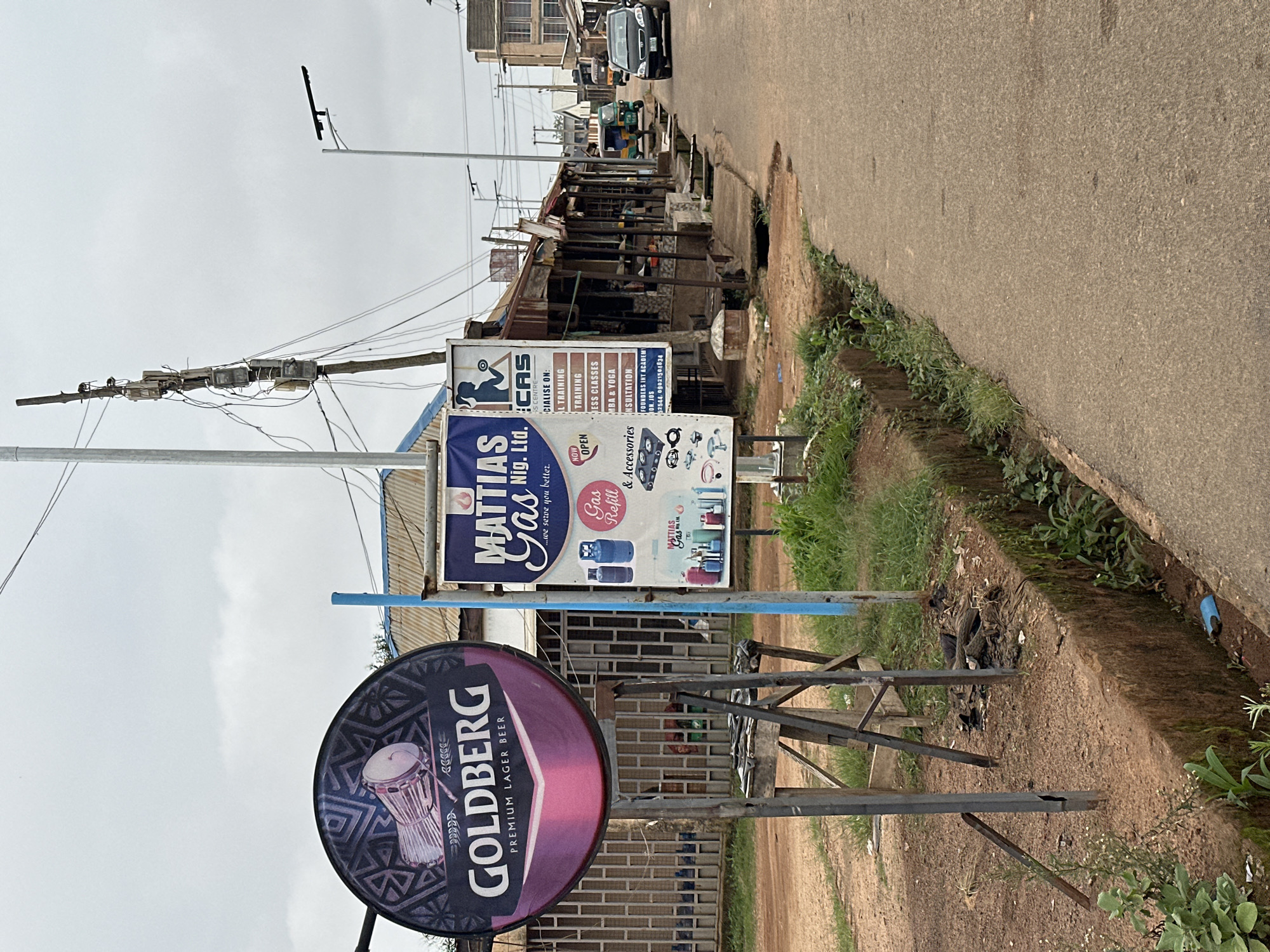 This is a medium shot, rotated 90 degrees counter-clockwise, of a street scene with commercial stalls and signs. The foreground features a circular sign for "GOLDBERG PREMIUM LAGER BEER" with a depiction of a drum. To its left, a vertical sign for "MATTIAS Gas Nig. Ltd." lists various services including "TRAINING," "ESS CLASSES," "MBA & YOGA," and "INSULTATION," with contact information for "FOUNDERS INT ACADEMY" in Jos. Behind these signs, a row of weathered market stalls with corrugated iron roofs lines the side of a paved road. The road is uneven and appears to have some potholes. Further down the street, a few vehicles are visible, including a car and a three-wheeled auto-rickshaw. The sky is overcast, suggesting a cloudy day. The scene depicts a typical street-level commercial area, likely in a developing country, with a focus on local businesses and their advertising. The overall impression is one of everyday commercial activity under a muted sky.