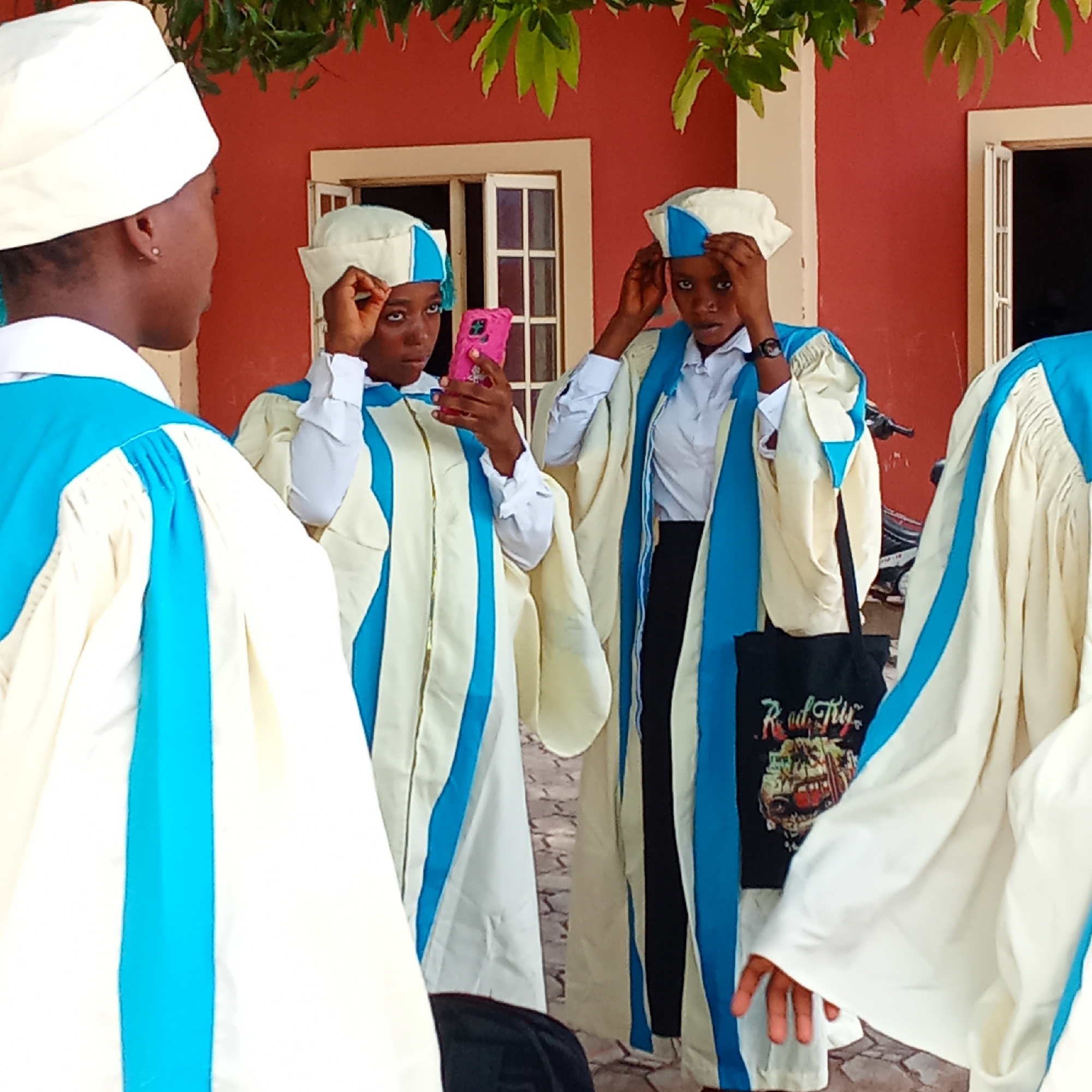 In front of a reddish-brown building with white-trimmed windows in an unknown city, a group of people wearing white and blue robes and hats are gathered. One person is holding a pink smartphone. The individuals appear to be adjusting their hats. A black tote bag is visible with a graphic design on it. Green leafy plants are visible at the top of the frame.