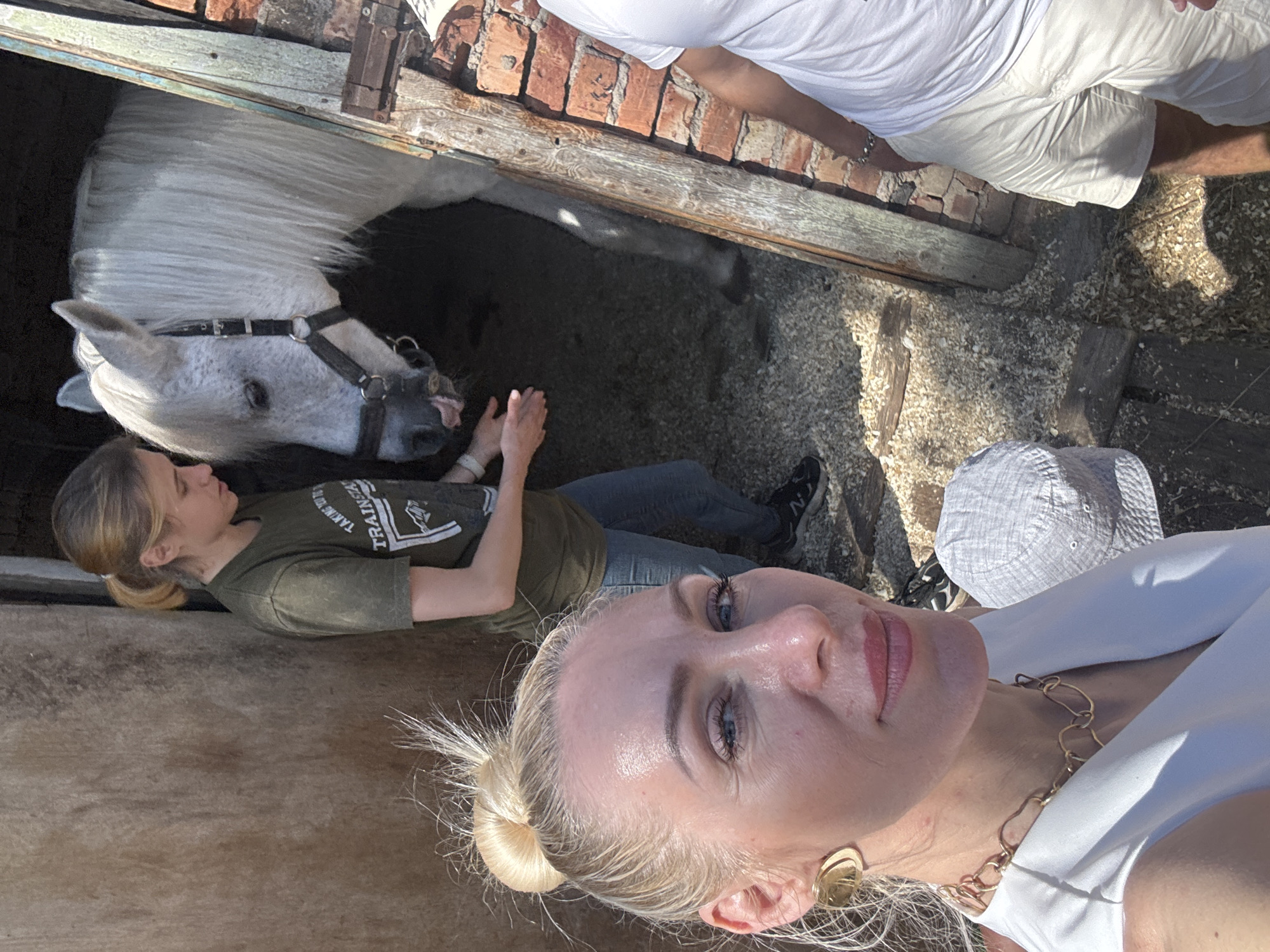 In Pavlovsk, Russia, a woman with blonde hair pulled into a bun, wearing a white top and gold jewelry, takes a selfie. Behind her, a younger woman in a green t-shirt and jeans stands near a white horse with a bridle, its tongue sticking out. The scene takes place in a rustic wooden and brick structure, possibly a stable, with wood shavings on the ground. Another person in a white t-shirt and light-colored shorts is partially visible on the right.