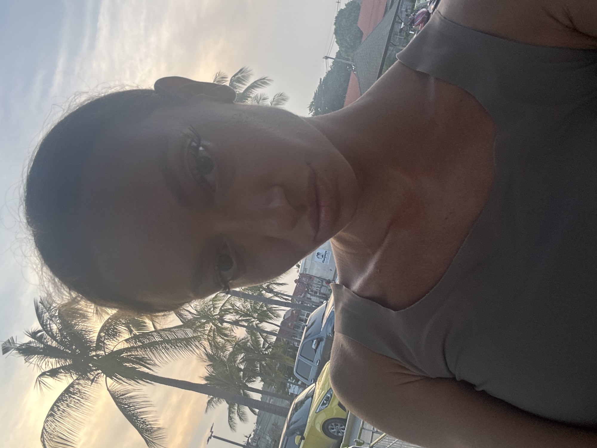 The image is a selfie of a woman taken outdoors in a tropical setting, consistent with Baan Bo Phut, Thailand. The woman is positioned in the foreground, looking towards the viewer with her head slightly tilted. Her face is partially shadowed, but her right eye (from the viewer's perspective) with visible eyelashes, her nose, and lips are discernible. She has dark hair and is wearing a light gray or tan sleeveless top.
In the background, numerous tall palm trees are prominent, silhouetted against a bright, hazy sky. The lighting suggests late afternoon or early evening, possibly sunset, as evidenced by the warm glow in the sky. Cars, including a yellow vehicle, are partially visible behind the palm trees, suggesting the location is a street or parking area. Faint outlines of buildings can be seen beyond the vehicles. The scene evokes a relaxed, tropical atmosphere. No specific activity is depicted beyond taking the selfie. No legible text is visible within the frame.