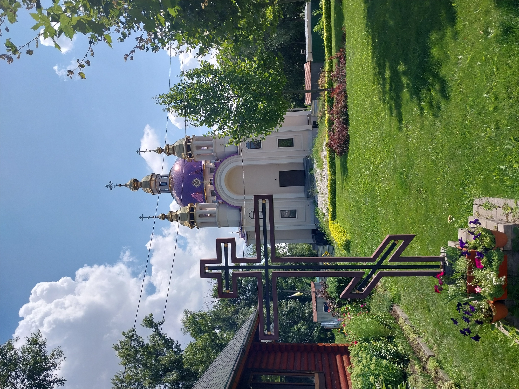 In Dnipro, Ukraine, a scene unfolds on a sunny day with scattered clouds. In the foreground, a large, stylized cross with burgundy and gold accents stands prominently in a grassy courtyard. Flower pots with purple and pink blooms are arranged at the base of the cross. To the left, a rustic wooden building with a slanted roof and a visible window is situated. Behind the cross and building, an ornate church with a large, purple dome and multiple gold-capped spires dominates the background. The church is white with subtle pastel accents and features arched windows and doorways. The surrounding environment is lush and green, with manicured lawns, vibrant flowerbeds, and mature trees. Power lines are visible crossing the sky, and in the distance, glimpses of other structures and vegetation can be seen.