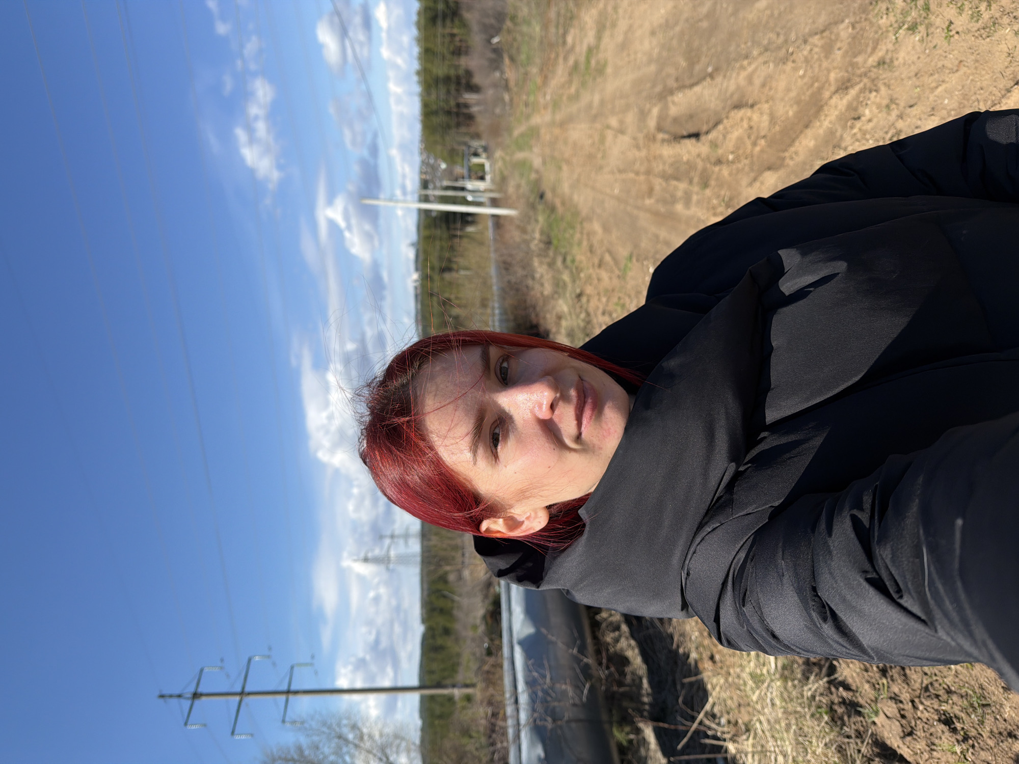 A close-up selfie, rotated 90 degrees counter-clockwise, features a young woman with shoulder-length red hair, fair skin, and light eyes, looking directly at the camera with a neutral expression. She is wearing a black puffer jacket and black pants.
The background reveals a bright, sunny outdoor scene under a clear blue sky dotted with scattered white clouds. To the right, a wide dirt or gravel path extends into the distance, with patches of dry grass and sparse vegetation. On the left and in the far distance, a line of trees forms a forest edge. Several tall utility poles carrying numerous power lines traverse the landscape, indicating a semi-rural or rural setting. A large, dark pipe or culvert runs horizontally across the mid-ground behind the woman. The bright daylight and the woman's warm jacket suggest a cool but sunny day, possibly in early spring or late autumn. There is no visible text in the image. The scene is located in Chaikovsky, Russia.