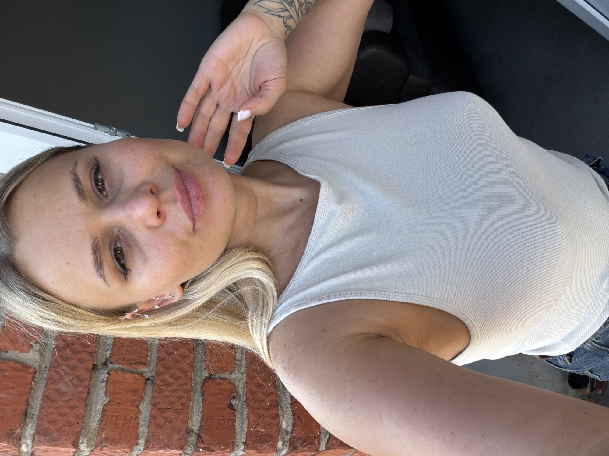 A woman with blonde hair is posing for a selfie. She is wearing a light-colored tank top. Her left hand is raised near her face, with white nail polish visible on her fingernails. She has multiple piercings in her left ear. Part of a brick wall is visible in the lower left corner of the image. The background also includes a window frame. The location is not discernable from the information provided.