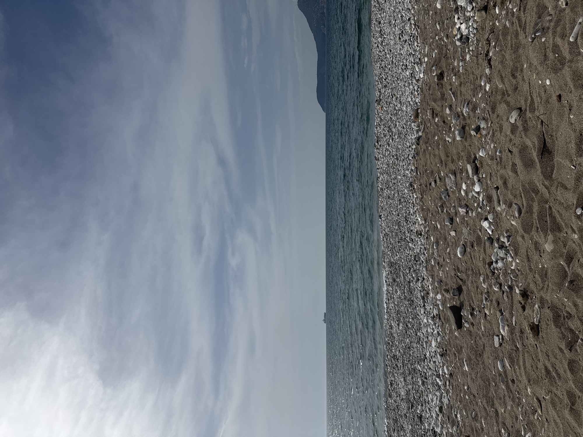 The image captures a serene coastal landscape in Bar, Montenegro, under an overcast sky. The scene is horizontally oriented, showing a beach, the sea, and a distant landmass.
In the foreground, a dark-sandy beach stretches, densely covered with countless small, light-colored pebbles and stones. The sand shows subtle ripples, indicating natural patterns. No people or objects are visible on the beach.
Moving towards the midground, the calm, blue-grey sea meets the shore. Gentle waves are visible, and sunlight subtly glints off the water's surface, creating small reflections.
The background features a large, dark landmass, likely a mountain or hill, rising from the sea on the left side of the frame, partially merging with the distant sky. On the horizon line, far out in the sea, a tiny, indistinct silhouette, possibly a distant ship or structure, can be observed.
The sky above is largely overcast with soft, diffuse clouds ranging from light blue-grey to white, suggesting a bright but not directly sunny day, likely late morning or early afternoon. The overall atmosphere is one of natural tranquility. There is no visible text in the image.