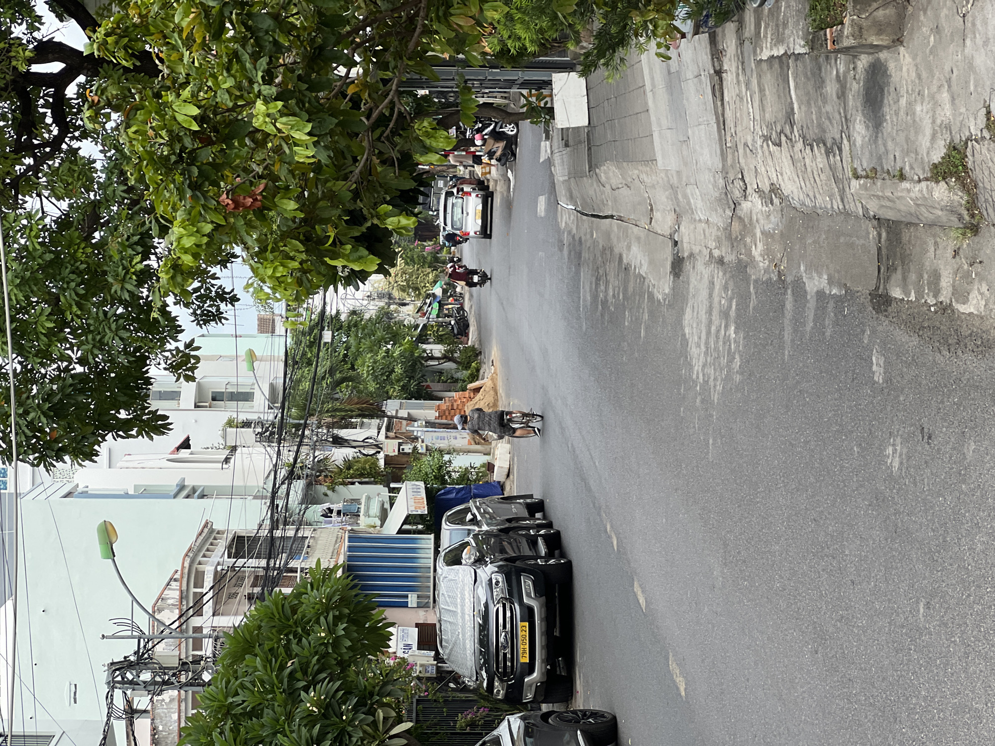 The image shows a street scene in Nha Trang, Vietnam. A black Ford SUV is parked on the left side of the street, its windshield covered with a silver reflective shade. Next to it, another SUV is parked. A man on a bicycle is riding away from the camera down the street. There are several buildings on the left side of the street, some with balconies and awnings. Trees and power lines are visible overhead. The street is paved and appears to be in a residential or commercial area. The overall impression is a typical urban street in Vietnam.