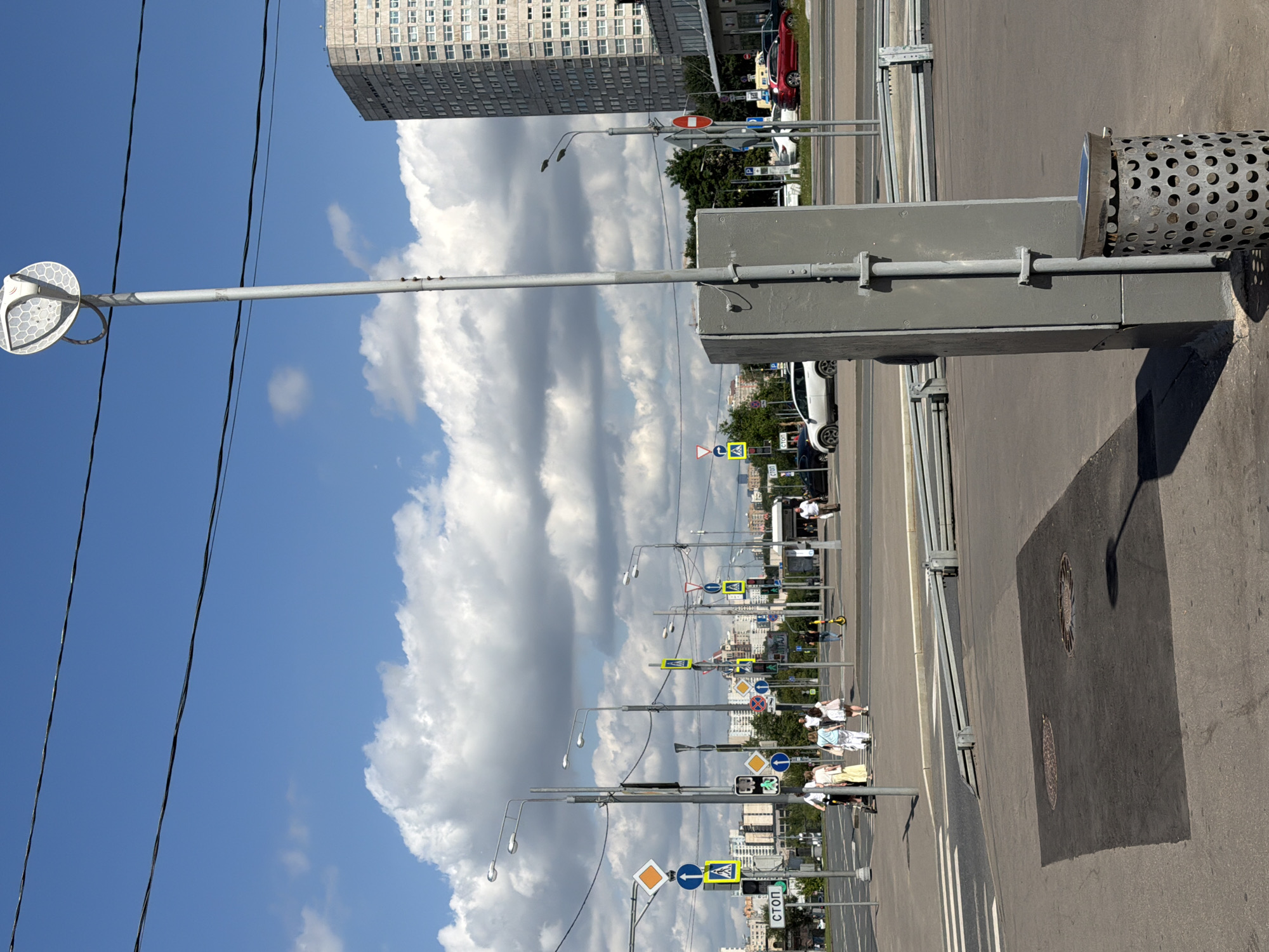 A bright, sunny day in Moscow, Russia, features a view of a city intersection with a large, modern building in the background. Several traffic signals and road signs are visible, including a pedestrian crossing sign and a stop sign. People are crossing the street, some on foot and others on scooters. A tall lamppost with a circular light fixture stands in the foreground, next to a grey utility box and a perforated metal trash can. The sky is blue with fluffy white clouds.