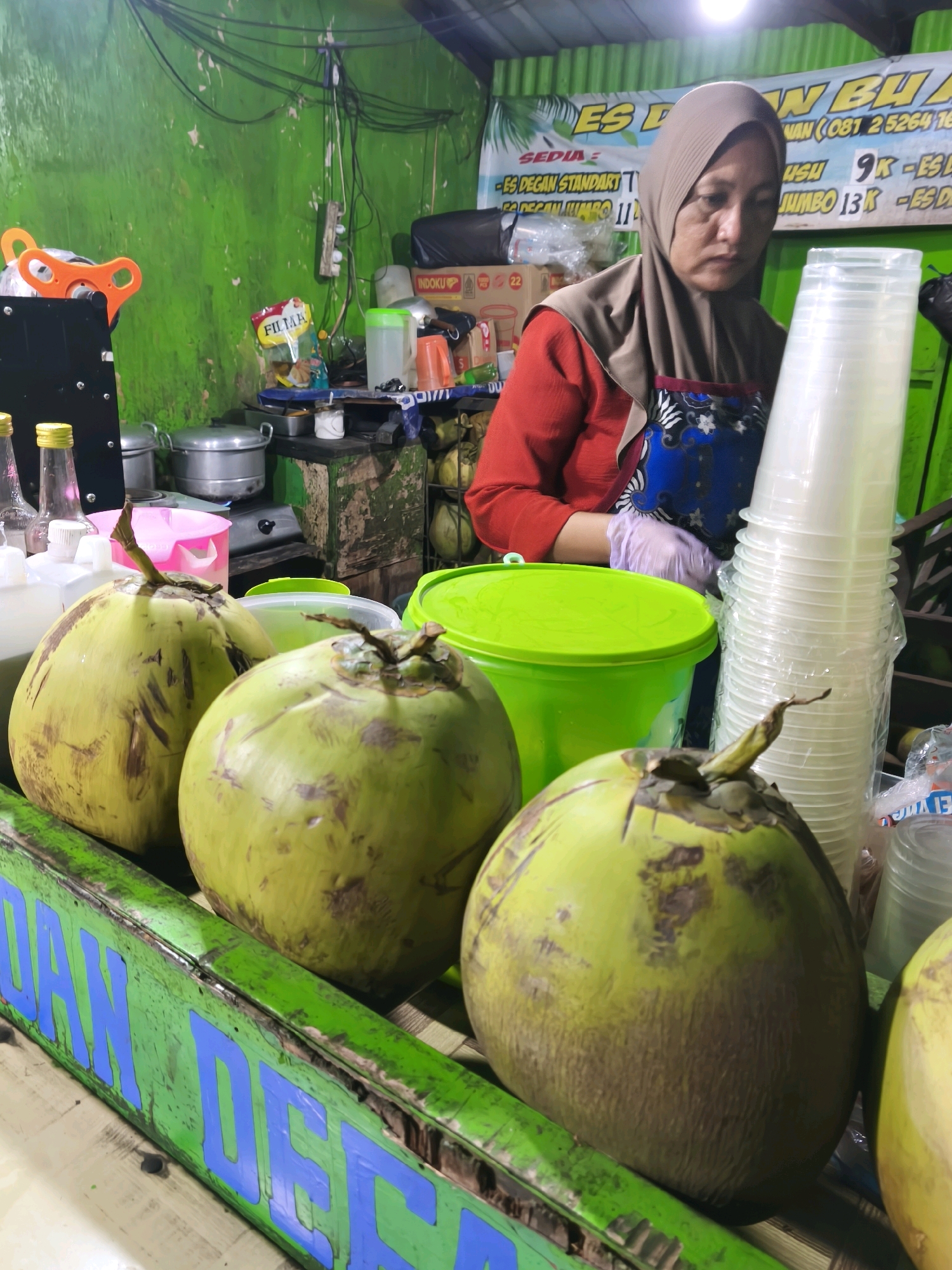 The image depicts a street-side stall in Surabaya, Indonesia, likely selling beverages. In the foreground, three large green coconuts are prominently displayed on a green wooden counter with blue lettering that appears to read "DAN DEAN". Behind the coconuts, a woman wearing a light brown hijab and a red shirt is visible. She is wearing purple gloves and appears to be preparing or serving something. To her right, a tall stack of clear plastic cups is packaged in plastic. Various items, including green plastic containers, stainless steel pots, bottles, and bags of snacks, are present on shelves and surfaces behind the woman, suggesting a commercial setting. The background wall is painted a vibrant green and shows some wear and tear, with electrical wires and a power outlet visible. The overall scene conveys a sense of a local, informal business operation in a tropical climate.
