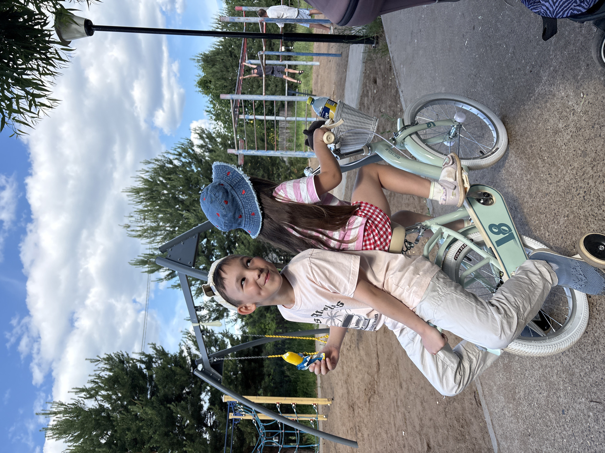 The image displays an outdoor scene at a playground in the city of Chaikovsky, Russia, on a day with a partly cloudy blue sky. In the foreground, two children are positioned on a light mint green child's bicycle equipped with white tires and training wheels. A boy, identified by a light-colored cap with prominent ear-like extensions, a pale short-sleeved t-shirt with a graphic, and light-colored pants, sits on the rear of the bicycle. He looks towards his right with a subtle smile, holding a yellow and blue toy in his right hand. A younger girl, wearing a blue denim bucket hat adorned with red hearts, a striped shirt, and a red and white checkered skirt, is seated on the main bicycle saddle, facing away from the viewer and gripping the handlebars. A small, light-colored wicker basket is attached to the front of the bicycle, containing a yellow plastic bottle and a dark object.
To the left of the children, a dark-colored metal swing set and a blue and yellow rope climbing structure are visible on a sandy playground surface, with tall pine trees in the background. To the right, a multi-colored metal monkey bar structure stands, with two individuals, appearing to be adults or older teens, engaged with it; one is hanging from the bars, and another stands nearby. A black lamppost with a light fixture is situated on the far right, partially obscured by green foliage. The children and bicycle are positioned on a dark paved path in the foreground.