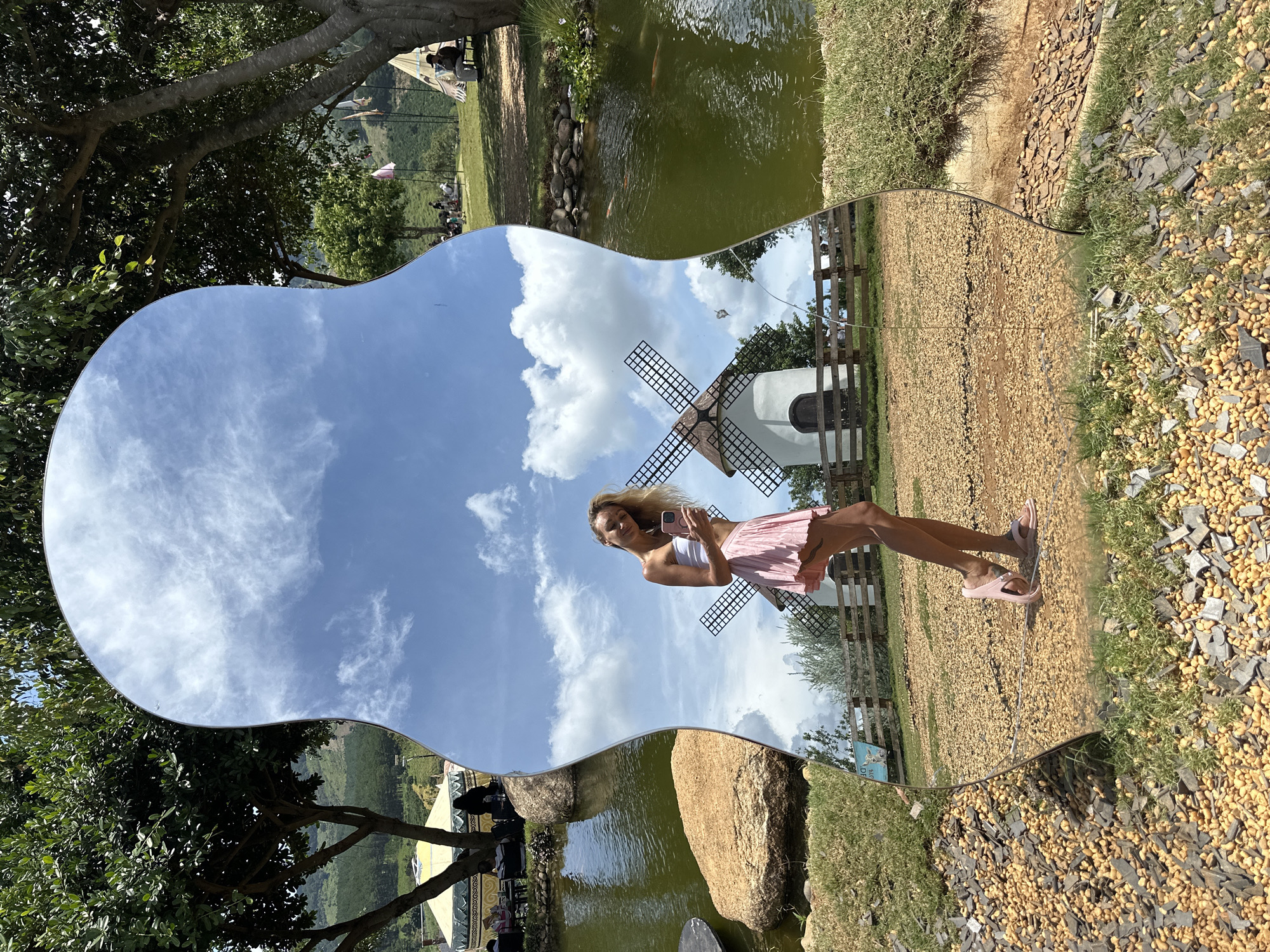 The image shows a woman in a white bandeau top and a pink pleated skirt posing for a selfie in a distorted mirror. She is holding a pink smartphone in her right hand and wearing pink sandals. The mirror reflects a scene with two white windmills, a wooden fence, and a clear blue sky with white clouds. In the background, there are trees, a pond with koi fish, and some tents. The foreground of the mirror shows a gravel path and grass. The location is identified as being in Thôn 5, Vietnam.