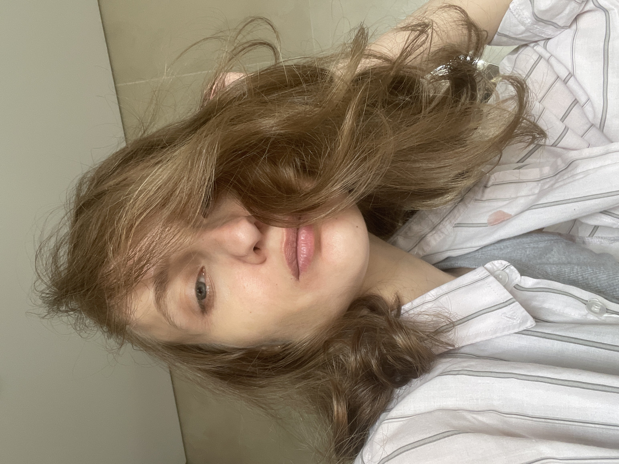 A fair-skinned person with light brown, wavy hair is the primary subject of this close-up, tilted portrait. The person is wearing a white button-down shirt with thin gray stripes. The background consists of a light-colored wall or surface and a tiled floor. There is no information available to determine if the person is located in Подшивалово, Russia.