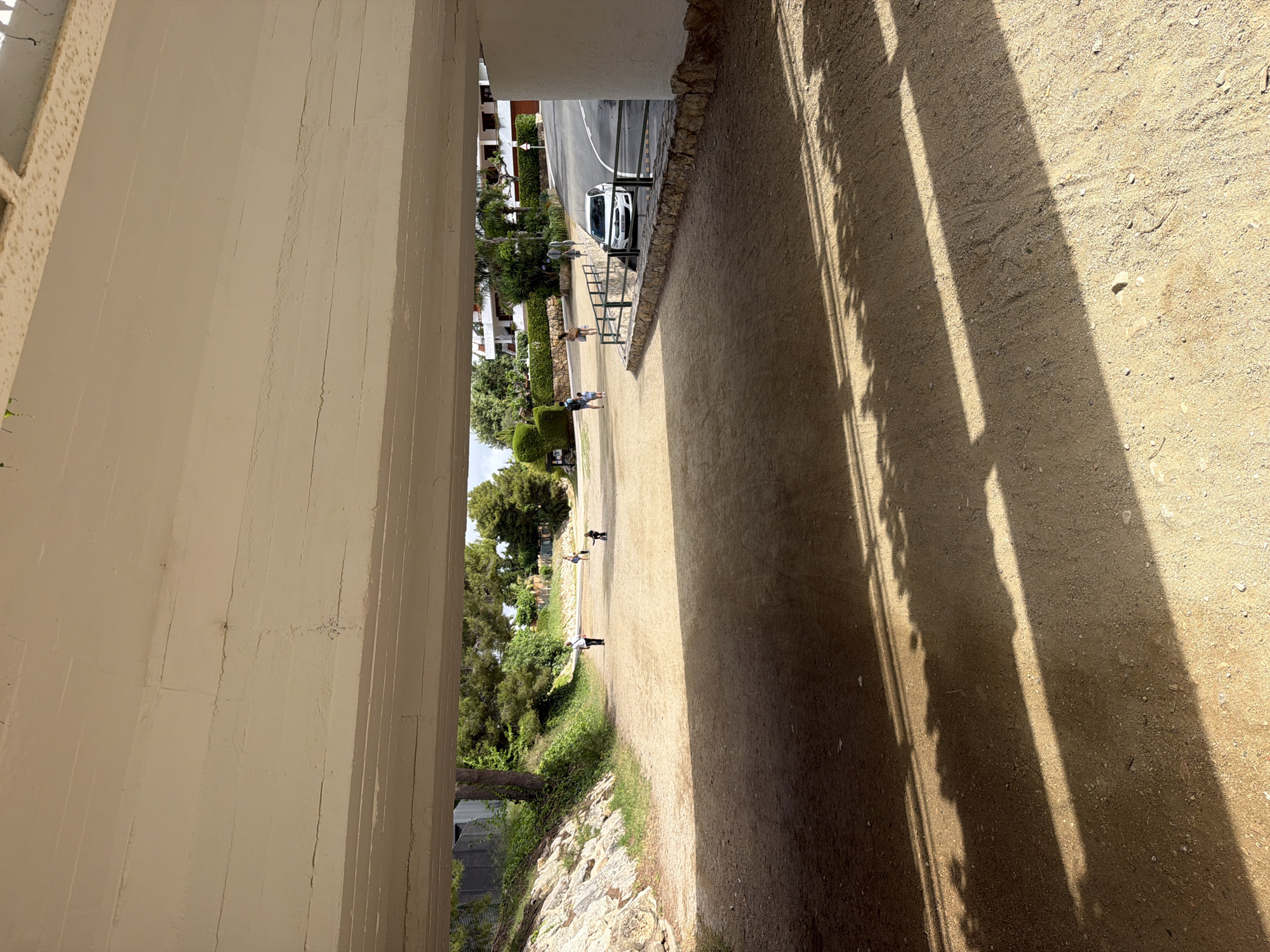 The image depicts an outdoor scene viewed from underneath a light-colored concrete overhead structure. The structure's underside shows horizontal lines, minor cracks, and subtle discoloration. The ground below is a wide, light brown dirt path or open area, with distinct elongated shadows cast by direct sunlight from the right.
Multiple individuals are present. Towards the far end of the dirt path on the left, an adult dressed in light attire walks rightward. In the central distance, a young child is seen on a balance bike, moving away from the viewer, accompanied by another adult figure. Further right on the path, another person walks away from the viewer. Adjacent to the paved road on the right, a group of three – two adults and a small child – is visible; one adult carries the child, while the other wears a light-colored dress.
A white compact hatchback car is parked on the right side of a paved, dark asphalt road that curves gently. The road is separated from the dirt path by a low stone wall topped with a dark metal fence. The car faces left, towards the underpass.
The left side of the scene beyond the dirt path features a rocky embankment covered with sparse green foliage, leading up to a dense cluster of various green trees, including pines. On the right side, beyond the road and fence, there are manicured green hedges and multi-story white buildings with reddish-brown tiled roofs, characteristic of Mediterranean architecture. The scene, located in Mont-roig del Camp, Spain, portrays a sunny day within a landscaped or residential area, suggesting a pedestrian underpass or access route.