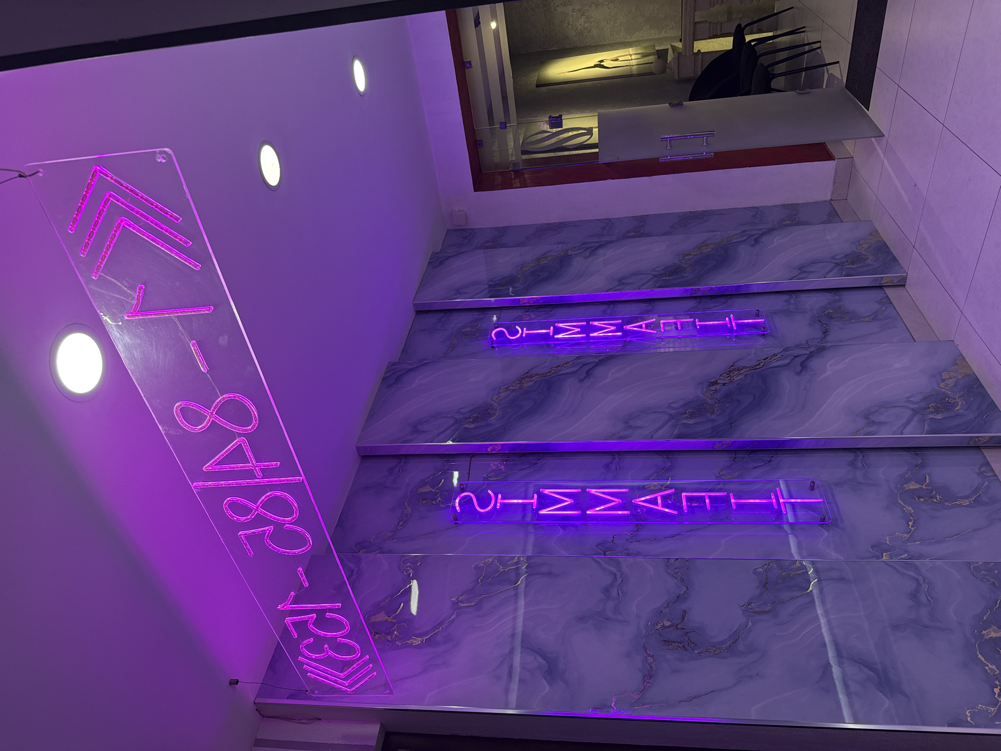 The image shows a modern interior hallway with purple neon lighting. A horizontal neon sign with Cyrillic text and arrows is mounted on the ceiling. Below, two vertical neon signs read "FITNESS" in Cyrillic script. The walls are decorated with a blue and purple marble-patterned laminate. To the right, a glass door leads to another room, where two black chairs are visible. The floor is tiled in white and gray squares. The overall impression is of a stylish and contemporary establishment, possibly a gym or spa. The presence of Cyrillic text suggests a location where this script is commonly used, such as Uzbekistan. Based on the visual cues and common architectural styles, this scene could be located in Tashkent, Uzbekistan.