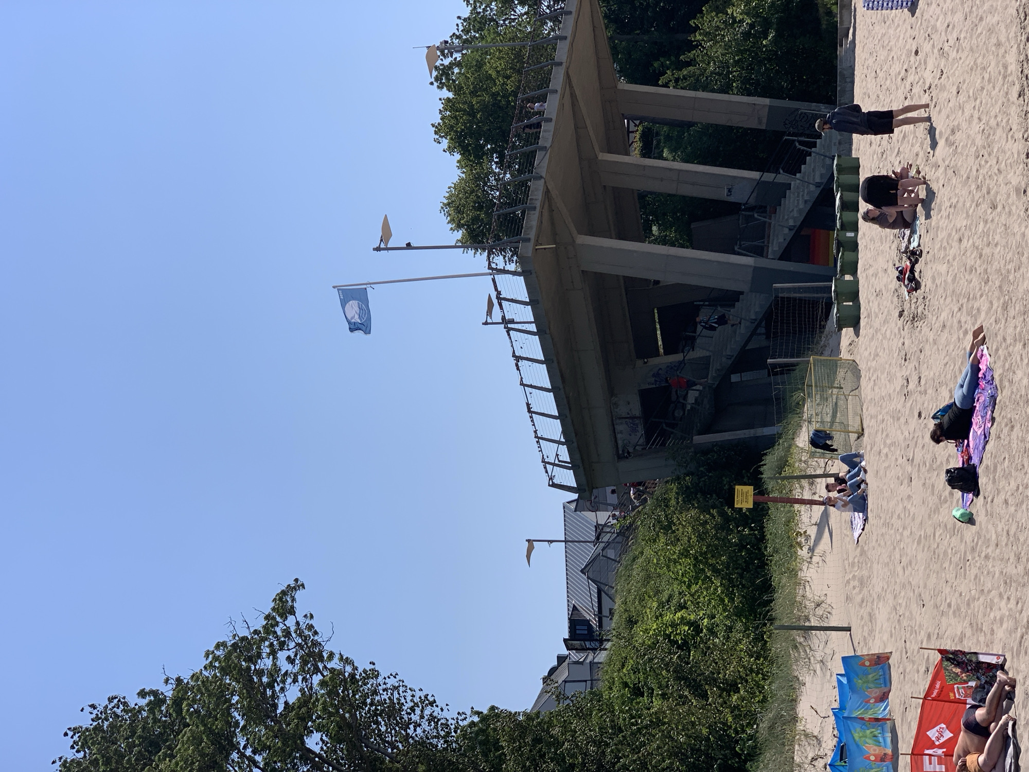 This is a daytime, eye-level, medium shot of a beach in Rewal, Poland. The sky is clear and blue, and the sun is shining. The foreground is occupied by a sandy beach with several people lounging and relaxing. Two people are sunbathing on a red windbreak in the lower left, while another person is lying on a purple towel. Further back, two people are sitting on a blanket, and a group of three people are gathered near a black backpack. There are also several green trash cans lined up.
In the middle ground, there are sand dunes and vegetation. A yellow sign is visible on a pole. Beyond the dunes, a large concrete structure with stairs and a walkway extends upwards. A blue flag with a white symbol is flying from a pole on the walkway. In the background, buildings with gray roofs and lush green trees are visible. The overall scene depicts a typical summer day at a beach resort.