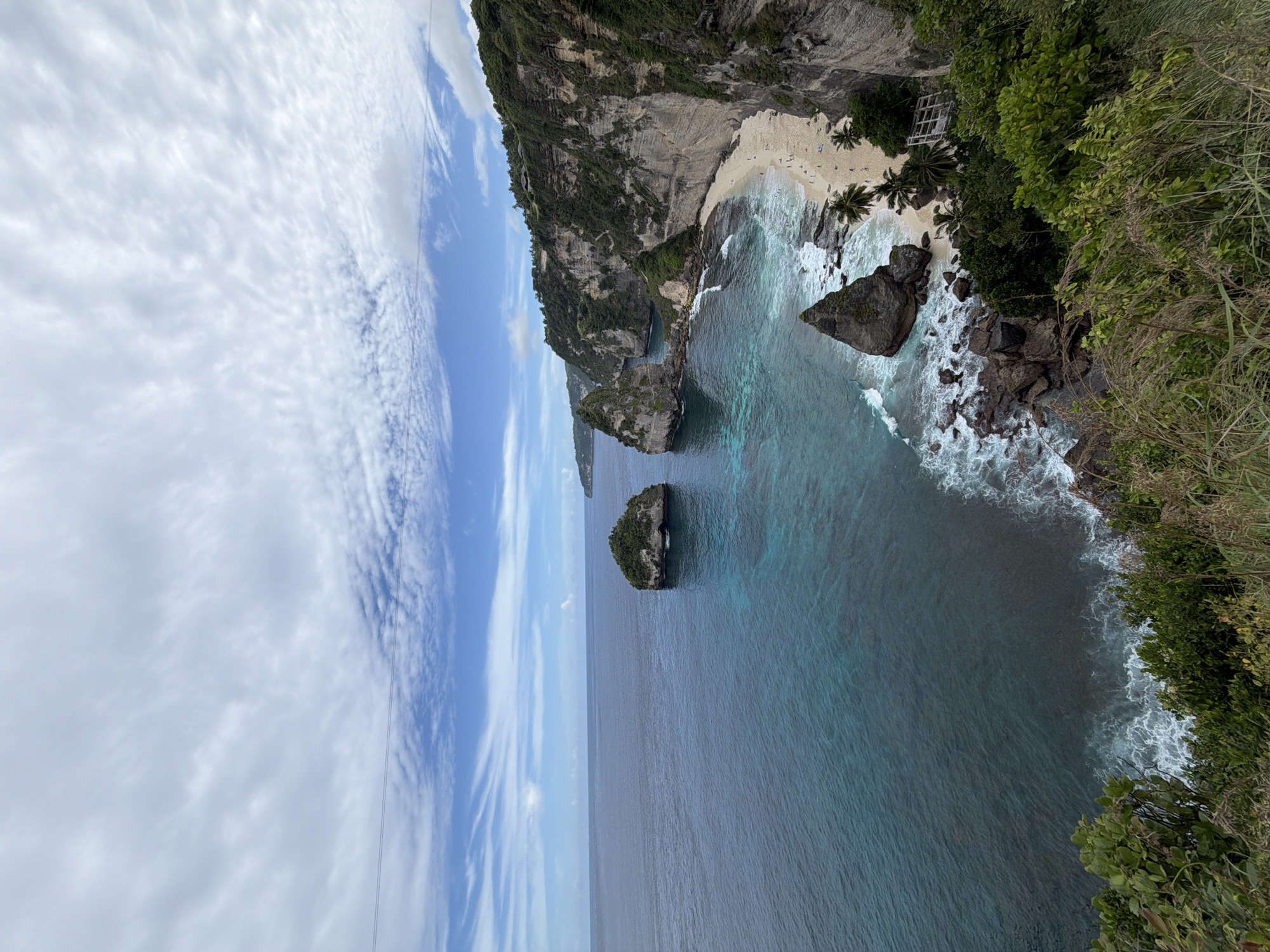 This landscape photograph presents a breathtaking, high-angle view of a tropical coastal scene in Nusa Penida, Indonesia. The vantage point is from a cliff edge, with green foliage and long grasses framing the immediate right foreground. The dominant feature on the right is a series of towering limestone cliffs, richly covered with vibrant green vegetation at their summits, descending sharply to meet the sea. Tucked into a serene cove at the base of these cliffs is a pristine white-sand beach, where several small figures, likely people, are visible. The beach is fringed with a few palm trees, and a light-colored, possibly wooden, structure is partially visible amidst the greenery near the shore.
The clear, turquoise waters lap against the sandy beach and large dark boulders scattered along the coastline, transitioning to a deeper, rich blue further out to sea. Several small, rocky islands or sea stacks punctuate the expansive ocean. The sky, filling the left side of the frame, is a canvas of bright blue with scattered, wispy white clouds that suggest fair weather. The scene is illuminated by bright daylight, indicating late morning or early afternoon. The overall impression is one of untouched natural beauty, showcasing the dramatic geological formations and crystal-clear waters characteristic of the region. There is no visible text in the image.