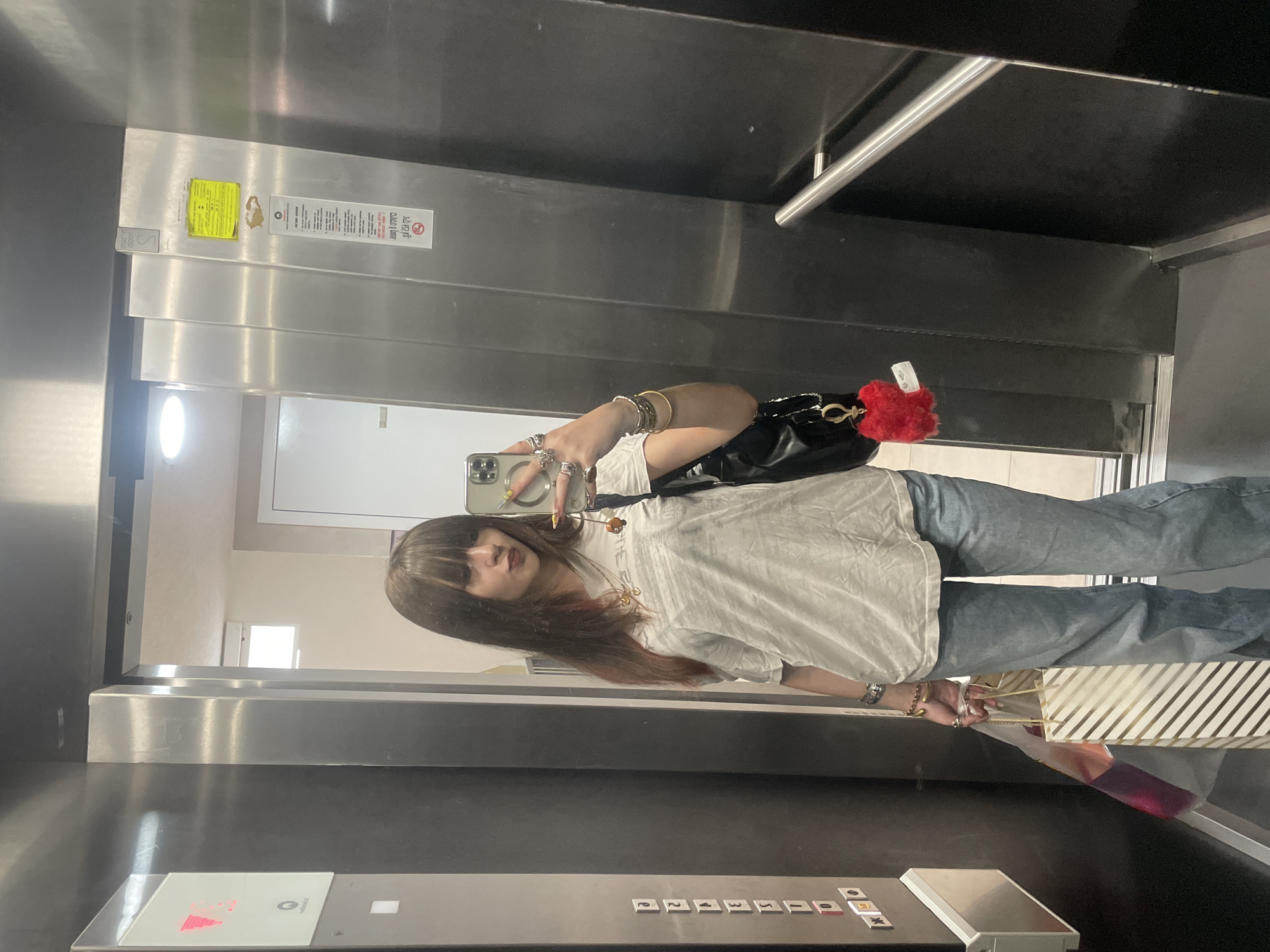 A young woman with long brown hair and bangs takes a mirror selfie in an elevator. She is wearing a white t-shirt, blue jeans, and multiple rings and bracelets. She is holding a shopping bag in her left hand and a black purse with a red fuzzy heart keychain is slung over her right shoulder. The elevator has metallic walls and buttons on the left side. A sign with Hebrew text is visible on the right wall. The location appears to be an elevator in Raanana, Israel, as indicated by the signage.