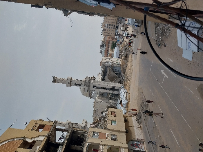 An elevated view captures a scene of widespread destruction in Khan Yunis, Palestinian Territory, centered around the heavily damaged Great Mosque of Khan Yunis. The mosque's tall, ornate minaret remains partially standing amidst significant structural collapse of the main building, which is reduced to rubble.
In the immediate left foreground, a multi-story residential building, possibly beige-colored, shows severe damage with large sections of its upper floors missing, exposing interior rooms and rebar. Laundry hangs from balconies on its lower, less damaged sections. On the ground floor, a faded poster is visible. To the far right, another partially destroyed building with exposed rebar and some hanging fabric frames the scene.
A paved street, marked with a white right-turn arrow, runs across the mid-ground. Approximately two dozen individuals, including men, women (some wearing hijabs), and children, are seen walking along the street and through areas heavily laden with debris. They move in various directions, navigating the devastated urban landscape.
Beyond the street and the mosque, the environment consists of extensive rubble piles, collapsed structures, and further damaged buildings. In the distance, a cluster of multi-story residential buildings, some appearing less damaged, forms the city's backdrop. Several vehicles, including a truck, are discernible in the far background. The sky above is a uniform light gray, indicating overcast conditions. The overall scene is one of post-conflict devastation, with inhabitants moving through the damaged environment.