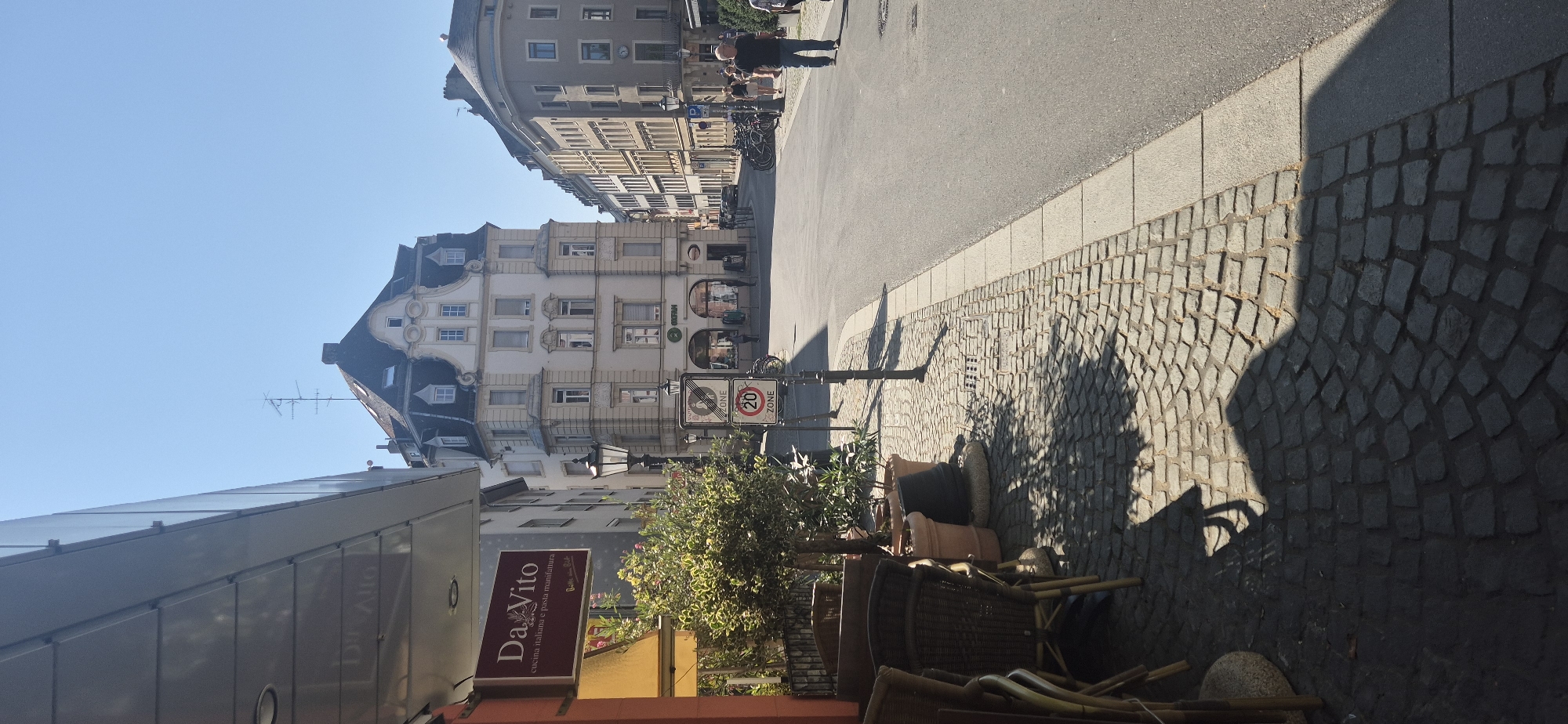 This is a street scene in Mainz, Germany, likely during daylight hours as indicated by the strong shadows. The perspective is from a sidewalk on the left, looking down a paved street.
On the left side of the image, there's a building with a modern, sleek facade, possibly a shop or restaurant, with a sign that reads "Da Vito" in red and white. This sign also indicates "cucina italiana e pasta manifattura." Below this overhang, outdoor seating with wicker chairs and tables is visible, along with potted plants. The sidewalk is paved with cobblestones.
Further down the street, a multi-story building with a light-colored facade and a mansard roof is prominent. It has several windows and architectural detailing. A street lamp stands on the sidewalk near the foreground. Traffic signs are visible, including one indicating a "ZONE" with a "20" speed limit.
Across the street, other buildings line the roadway, some appearing to be older architecture with multiple stories and varying facade colors. A row of parked bicycles is visible against one of the buildings.
Several people are present on the street. In the middle distance, a person in a dark shirt and blue jeans walks away from the camera. In the background, other figures are sparsely distributed on the sidewalks and street. The sky is clear and blue. The overall atmosphere suggests a European city street on a sunny day, with commercial establishments and pedestrian activity.