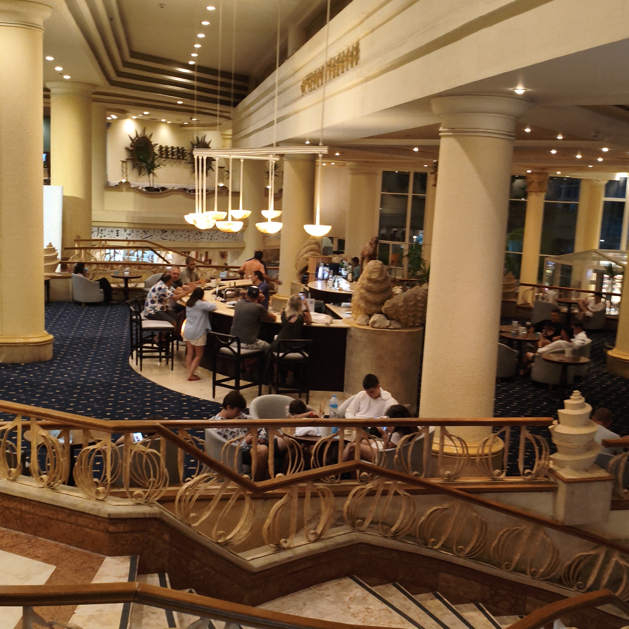The image depicts the interior of a hotel lobby or lounge area. The scene is well-lit by a combination of ambient ceiling lights and decorative pendant lamps. Several people are visible, engaged in various activities.
In the foreground, a grand staircase with ornate wooden railings leads down. On the landing of the staircase, several individuals are seated in chairs, appearing to be relaxing or interacting with their mobile devices.
Further into the lounge, a bar area is prominent. Patrons are seated at the bar and at small tables, with some ordering drinks and others conversing. A few staff members are behind the bar, serving customers. The decor features large, cream-colored columns, dark blue patterned carpeting, and decorative wall accents. Sculptural elements, possibly resembling animals or abstract forms, are placed near the bar. The overall atmosphere suggests a social gathering or a casual evening within the hotel.
The lighting and the attire of the people suggest it is evening. There are no clear indications of the specific location or any weather conditions. No visible text is present in the image.