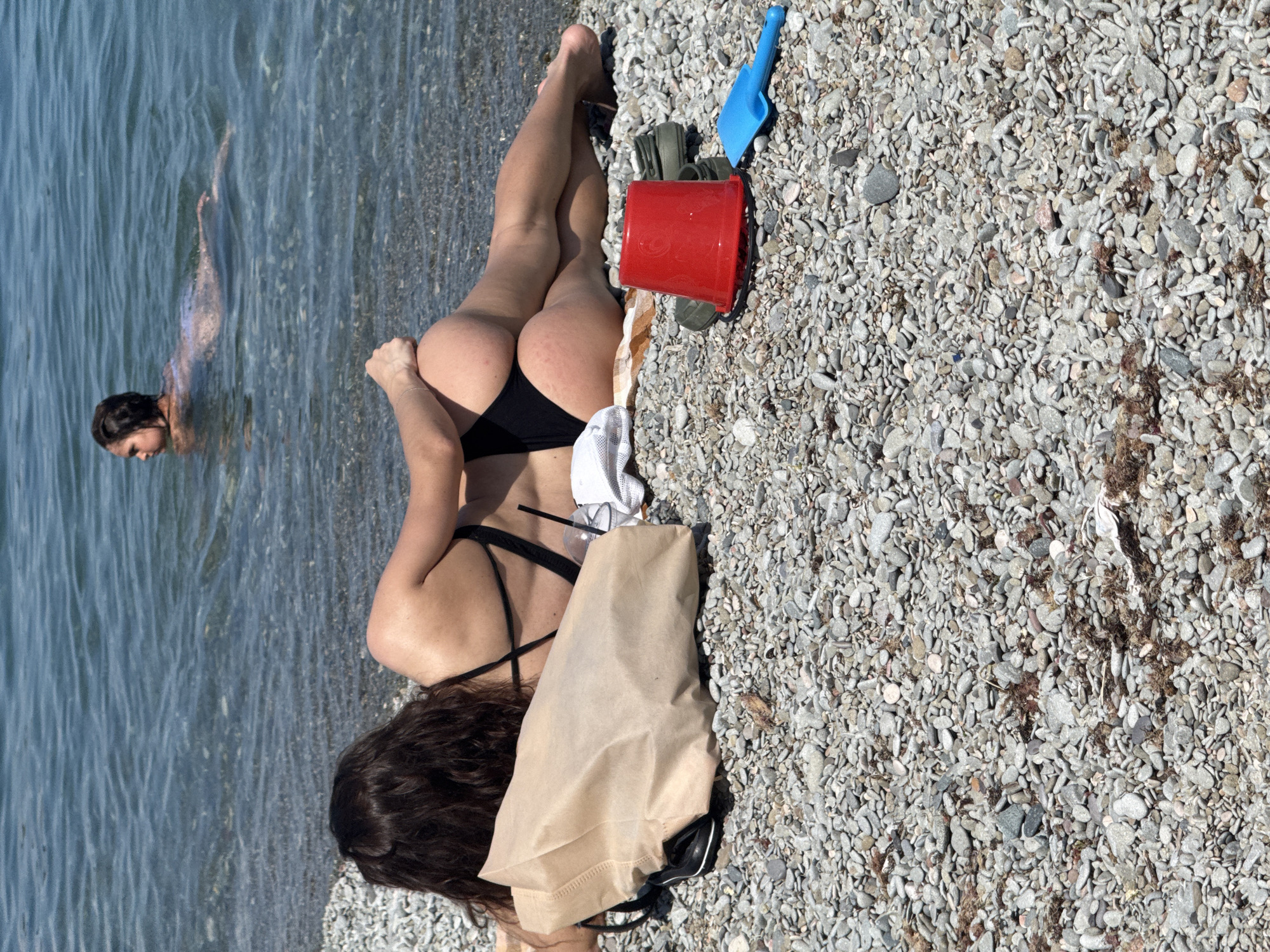 A high-angle, outdoor daytime photograph shows a woman in a black bikini lying on a rocky beach, while another woman swims in the sea. The woman lying on the beach has dark, curly hair, fair skin, and is positioned on her stomach with her legs facing the upper left of the frame. She has a brown paper bag, a white towel, a red bucket, and a blue shovel nearby. The beach is composed of small gray pebbles. In the upper left of the frame, another woman with short, dark hair swims in the blue sea.
