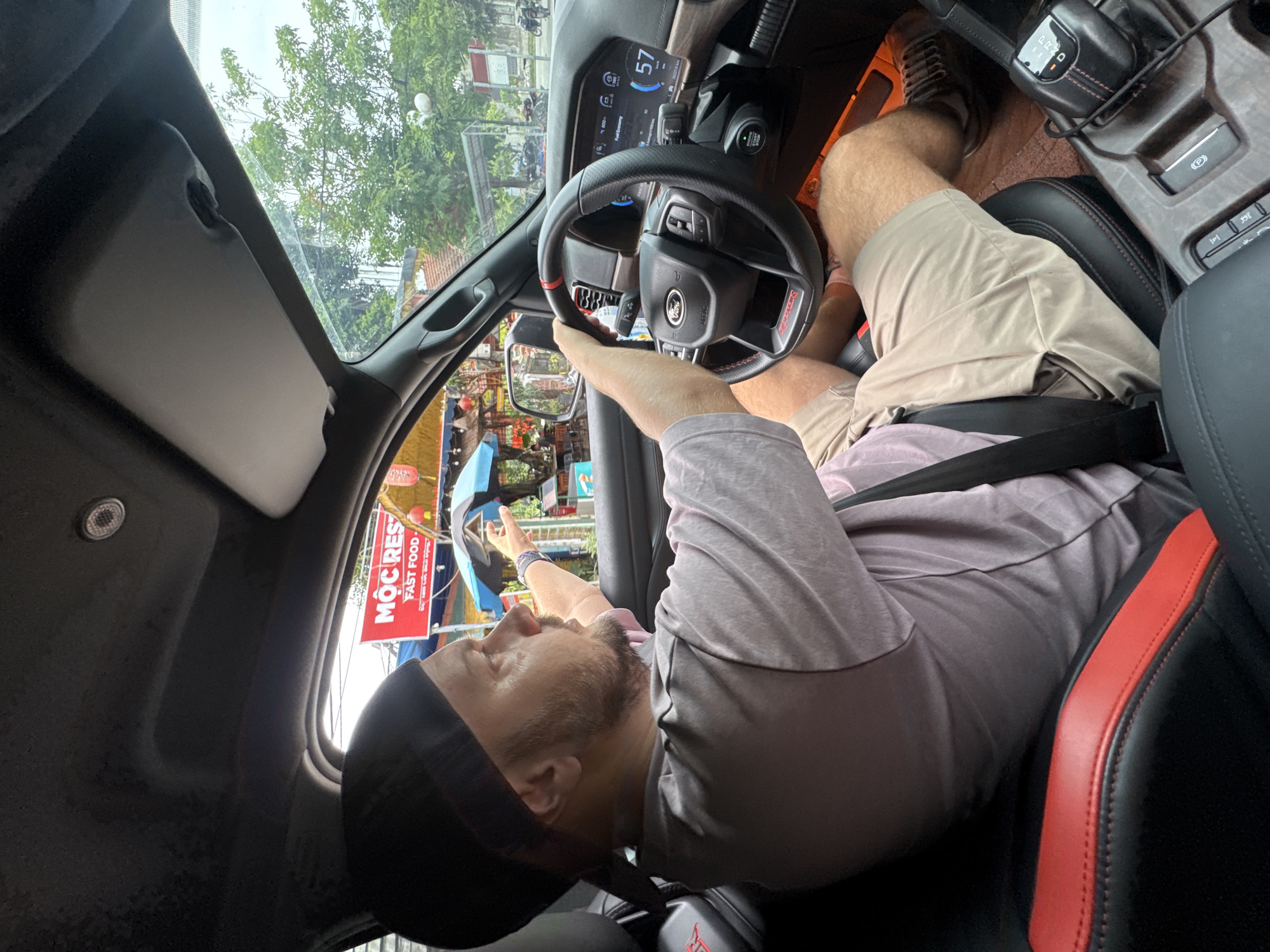 In Nam Hoa Lư Ward, Vietnam, a man wearing a black skullcap and a light purple t-shirt is seated in the driver's seat of a Ford vehicle, holding the steering wheel. He is pointing his left hand outward, seemingly indicating something outside the car. The car's interior features black seats with red accents, a digital dashboard displaying a speed of 57 km/h, and a gear shifter. Outside the car, a sign for "MOC REST. FAST FOOD" is visible, along with trees and other structures. The scene appears to be an urban or suburban environment.