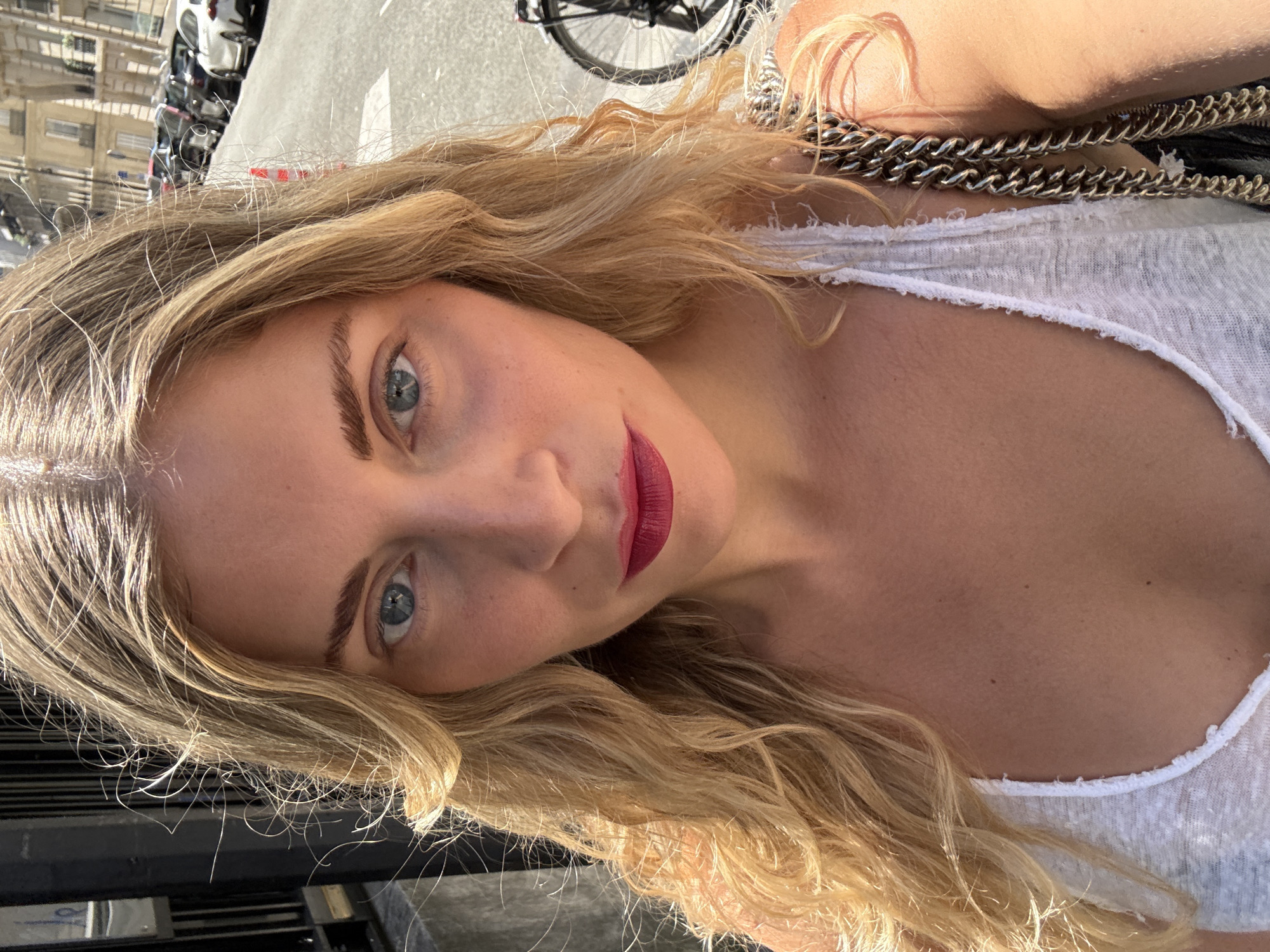 A fair-skinned woman with long, wavy blonde hair and blue eyes is captured in a close-up photograph, tilted at an angle. She is wearing a light-colored, possibly white, low-cut shirt or tank top. Her lips are painted with a bold, dark red lipstick. A chain strap from a bag is visible over her shoulder.
The background is slightly blurred, but contains visual information indicative of Paris, France. Reflections of buildings, including those with ornate balconies, are visible. Parked cars and motorcycles are also apparent, typical of urban street scenes in Paris. A bicycle is partially visible in the background. The scene suggests an outdoor setting during daylight hours, likely on a street.
