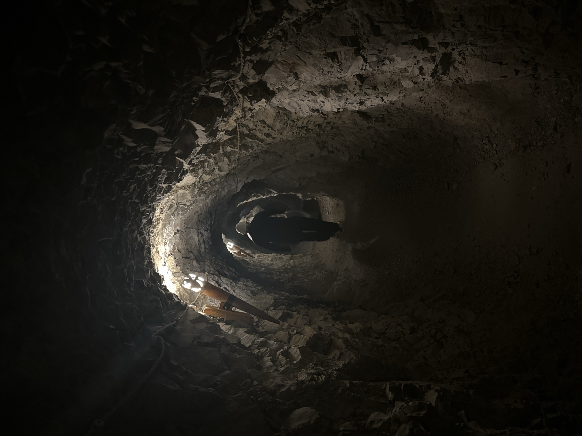 The image depicts an underground tunnel in the city of Novo-Zubutli, Russia. The tunnel features rugged, uneven rock walls and a dirt pathway. Artificial light fixtures, resembling stylized torches or lamps, are mounted at intervals along the left wall, casting illumination forward into the dark passage. A single individual, viewed from behind, walks deeper into the tunnel, wearing dark clothing and a light-colored hard hat that is brightly lit. A rope handrail is visible along the left wall. The environment is characterized by its confined, rocky structure, suggesting a mine shaft or a prepared cave passage.