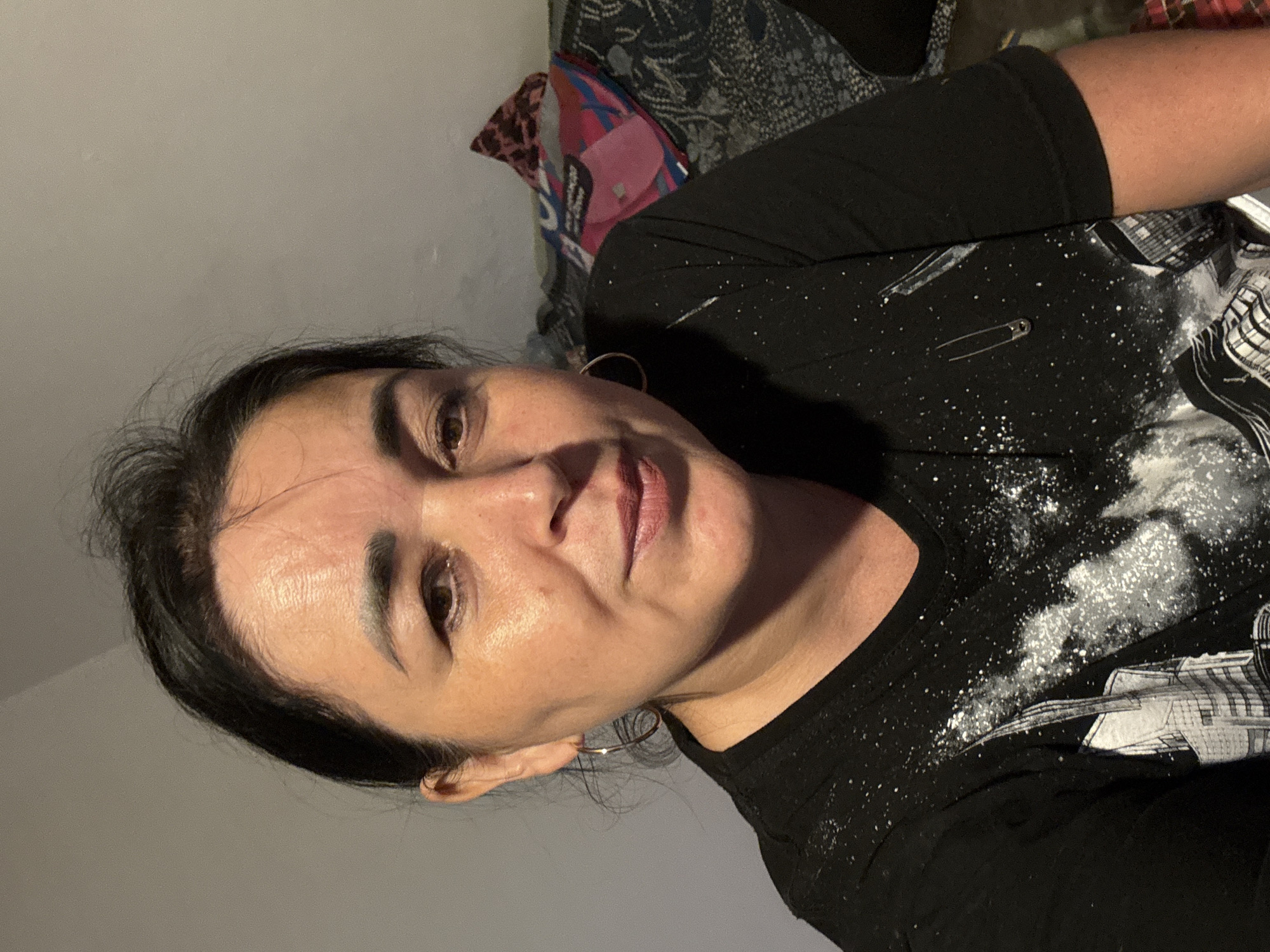The image is a portrait of a woman in Starokhalilovo, Russia. She is looking towards the camera with her head tilted. The woman has dark hair and is wearing a black t-shirt with a white speckled pattern and a graphic design. She is also wearing hoop earrings. The background is blurred, but it appears to be an indoor setting with a gray wall and some patterned fabric. A small safety pin is visible on her shirt. The lighting suggests it is either daytime or artificial indoor lighting. The woman's expression is neutral.