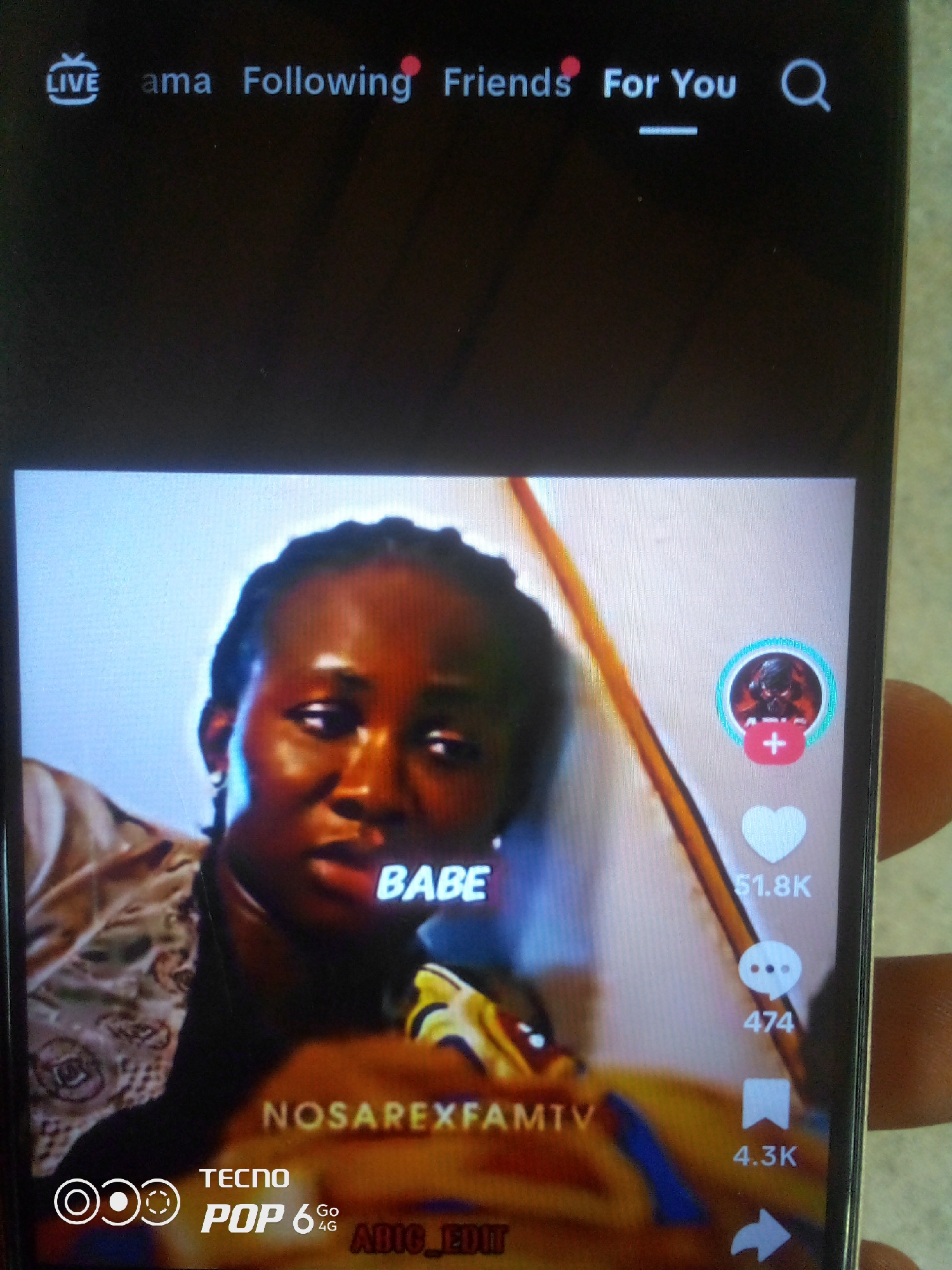 The image shows a close-up of a smartphone screen displaying a video. The top of the screen has a navigation bar with options: "LIVE," "ama," "Following," "Friends," "For You," and a search icon. The video content features a woman with braided hair looking towards the viewer. Text overlays on the video include "BABE" near the woman's mouth, "NOSAREXFAMTV" in the lower center, and "TECNO POP 6 4G ABIG EDIT" in the bottom left corner. On the right side of the screen, there are icons for user interaction: a profile picture with a red plus sign, a heart icon with "51.8K" below it, a speech bubble icon with "474" below it, and a bookmark icon with "4.3K" below it. A finger is visible on the right edge of the phone, holding it. The background of the video appears to be a plain, light-colored wall. There is no visible indication of the city of Howayi, Nigeria, in the image.