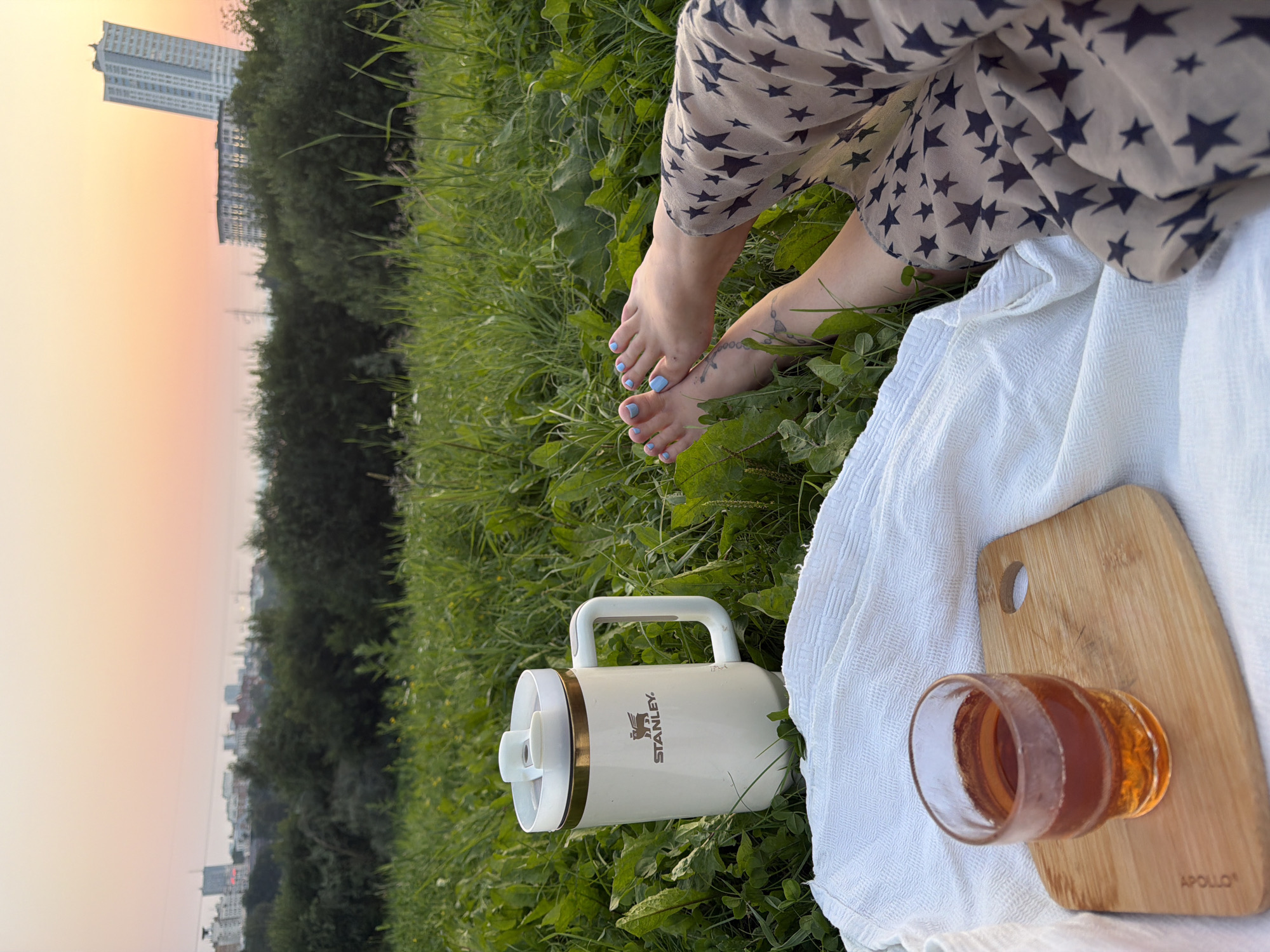 An adult's bare feet, with blue nail polish, rest on a picnic blanket spread across a grassy field. The person is wearing a tan dress with a dark star pattern. To the left, a white and gold insulated mug with the "STANLEY" logo sits in the grass. Next to the mug, a glass of amber liquid rests on a wooden cutting board, which is also on the blanket. In the background, a tree line separates the field from a city skyline, characterized by tall buildings and a distinct, slender skyscraper. The sky is a soft gradient of pale orange and pink, indicating either sunrise or sunset. This scene is consistent with the outskirts of Perm, Russia, given the architecture and natural landscape.